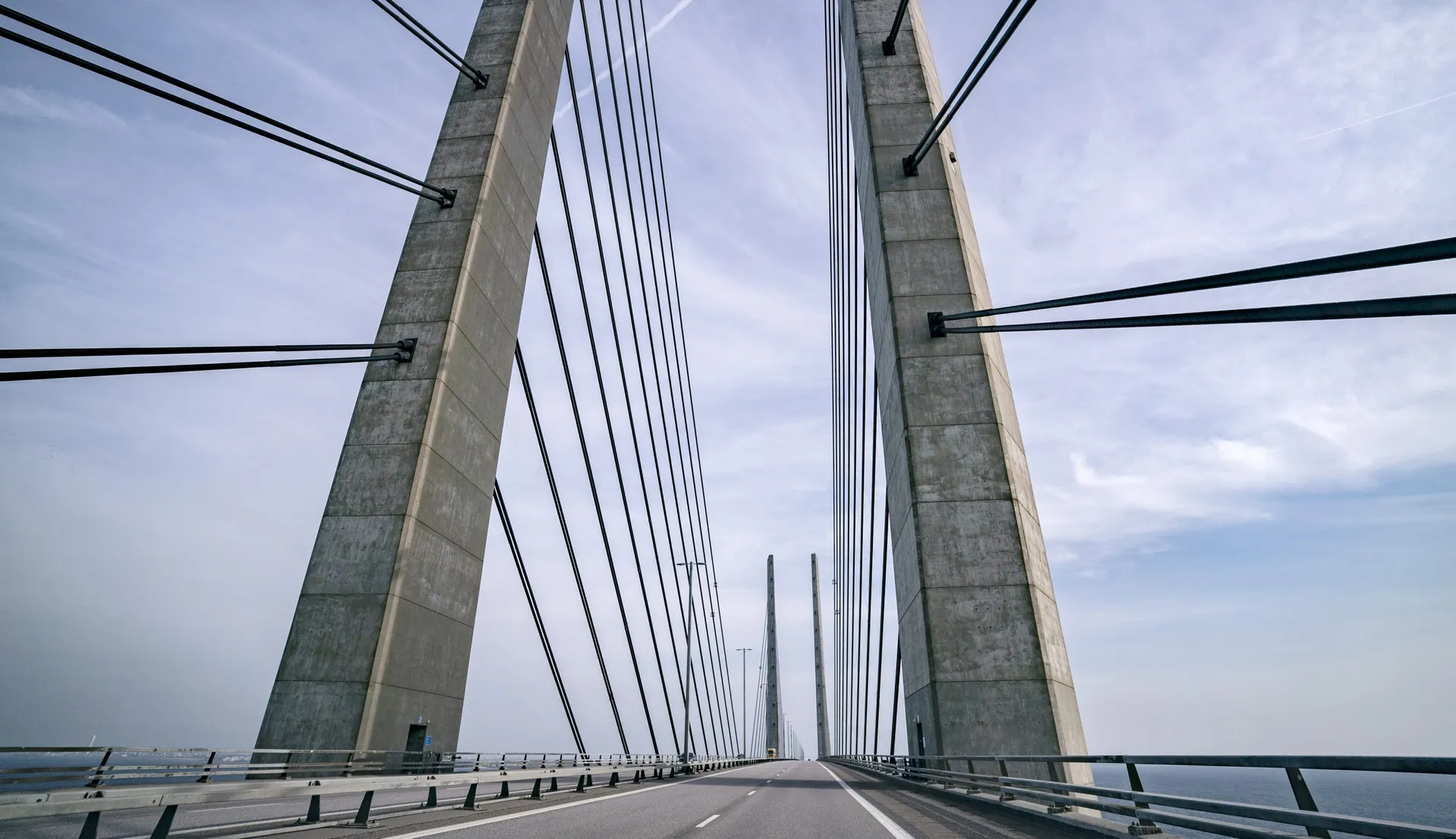 The image shows a large cable-stayed bridge with tall concrete pylons and numerous cables supporting the structure. The photo is taken from the roadway perspective, illustrating the symmetry of the bridge's design. The sky is clear with a few clouds.