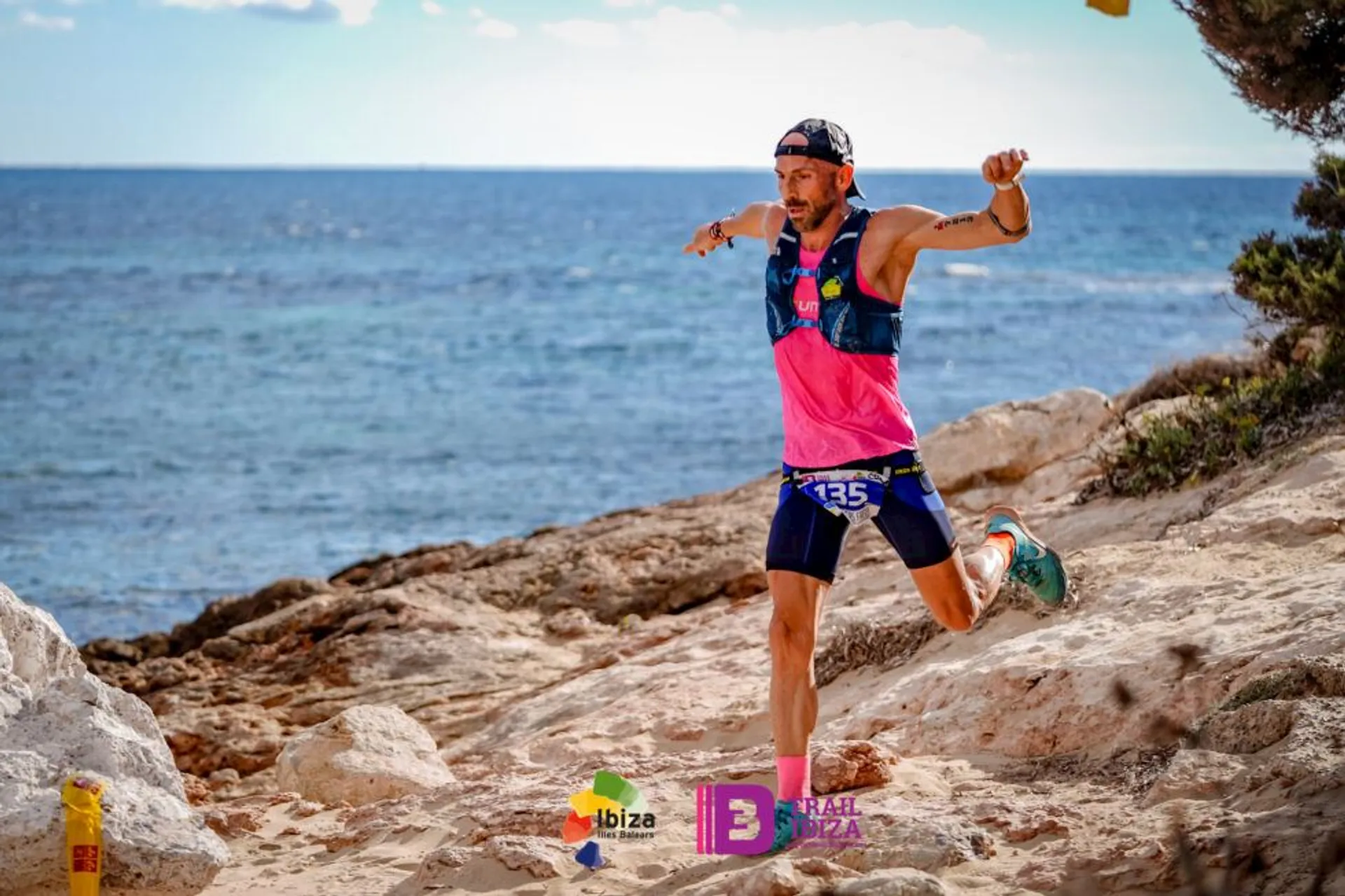 The image shows a person running on a rocky trail near the ocean, likely participating in a trail running event. The runner is wearing athletic gear, including a pink and blue outfit, and appears to be in motion with arms outstretched. The background features the sea and a clear sky. Various logos, including "Ibiza" and "Trail Ibiza," are visible at the bottom of the image, suggesting the location or event name.