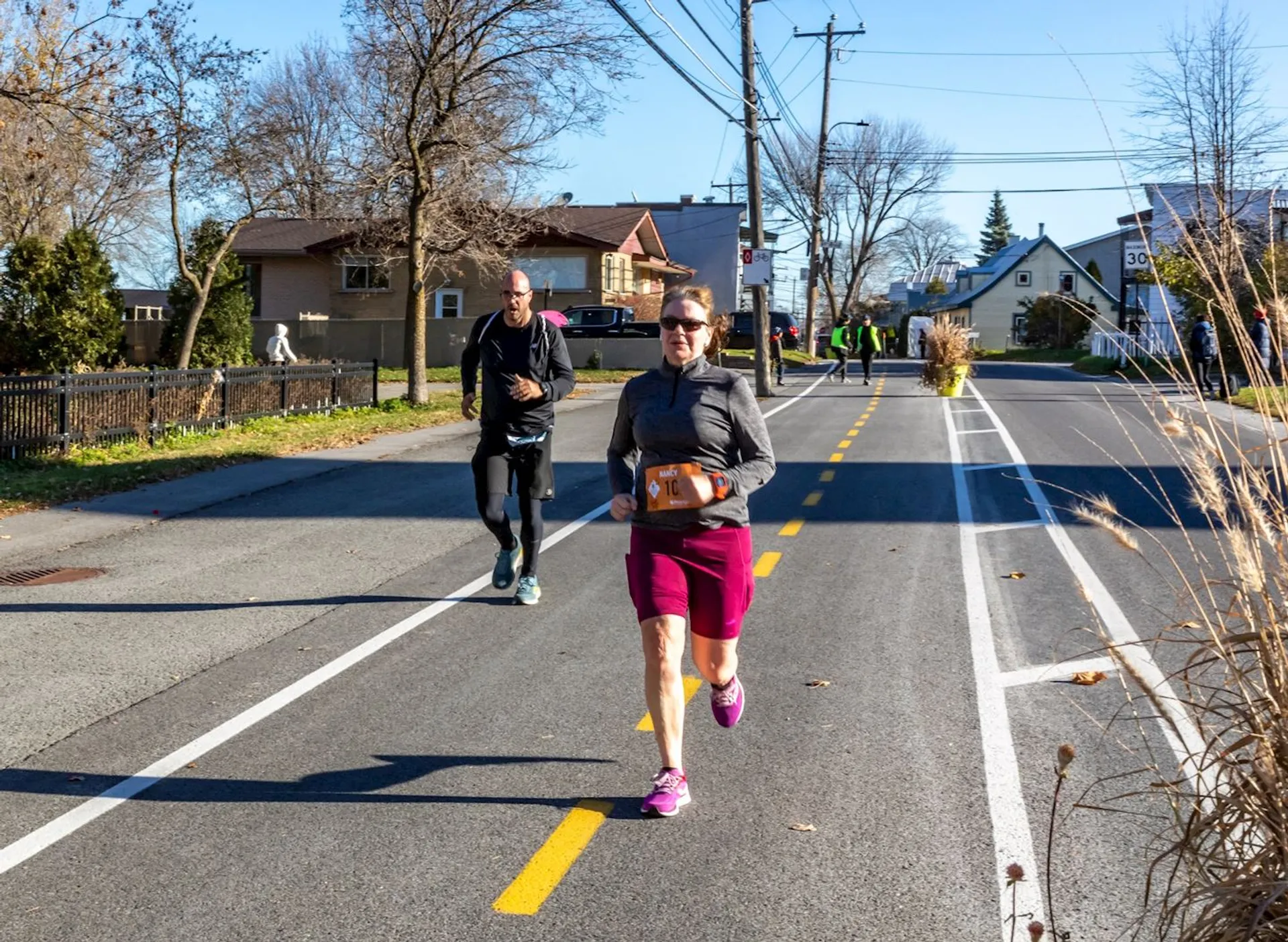 The image shows people running on a paved street, possibly participating in a running event. There are houses and trees along the road, and the weather appears to be clear and sunny.