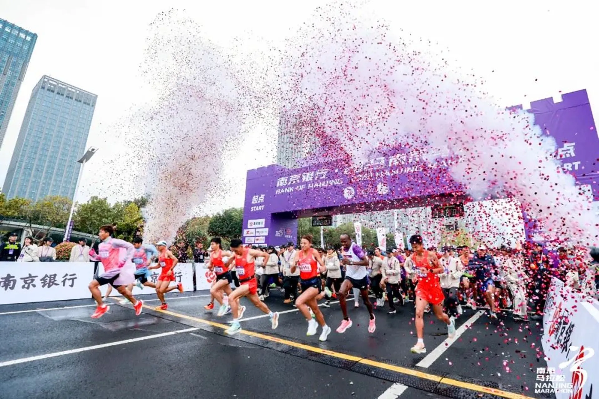 This image shows a group of runners at the start of a marathon or race event. There is confetti in the air, and a start line banner above them. People in the background are cheering, and some buildings are visible.