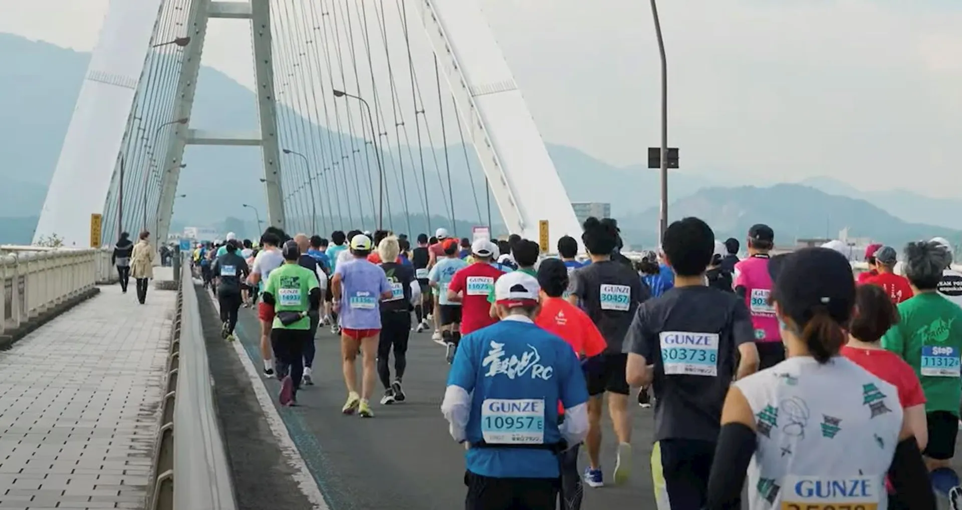 The image depicts a group of runners participating in a road race, likely a marathon or half-marathon, as suggested by the participant numbers visible on their running attire. They are running across a bridge with a distinctive arch structure, which lends a scenic backdrop to the event. The participants are dressed in typical athletic gear appropriate for a running event. The weather appears to be overcast, which can provide cooler conditions ideal for such endurance events.