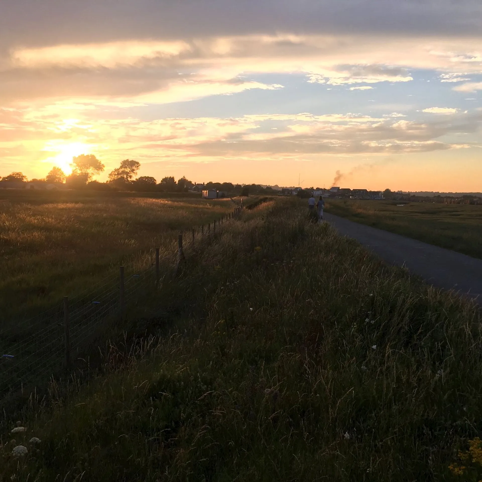 This image depicts a scenic landscape at sunset. It features a path alongside grassy fields with a fence. The sky is filled with a mix of clouds and the warm hues of a setting sun. There are people walking on the path in the distance and silhouetted trees and buildings can be seen against the horizon.