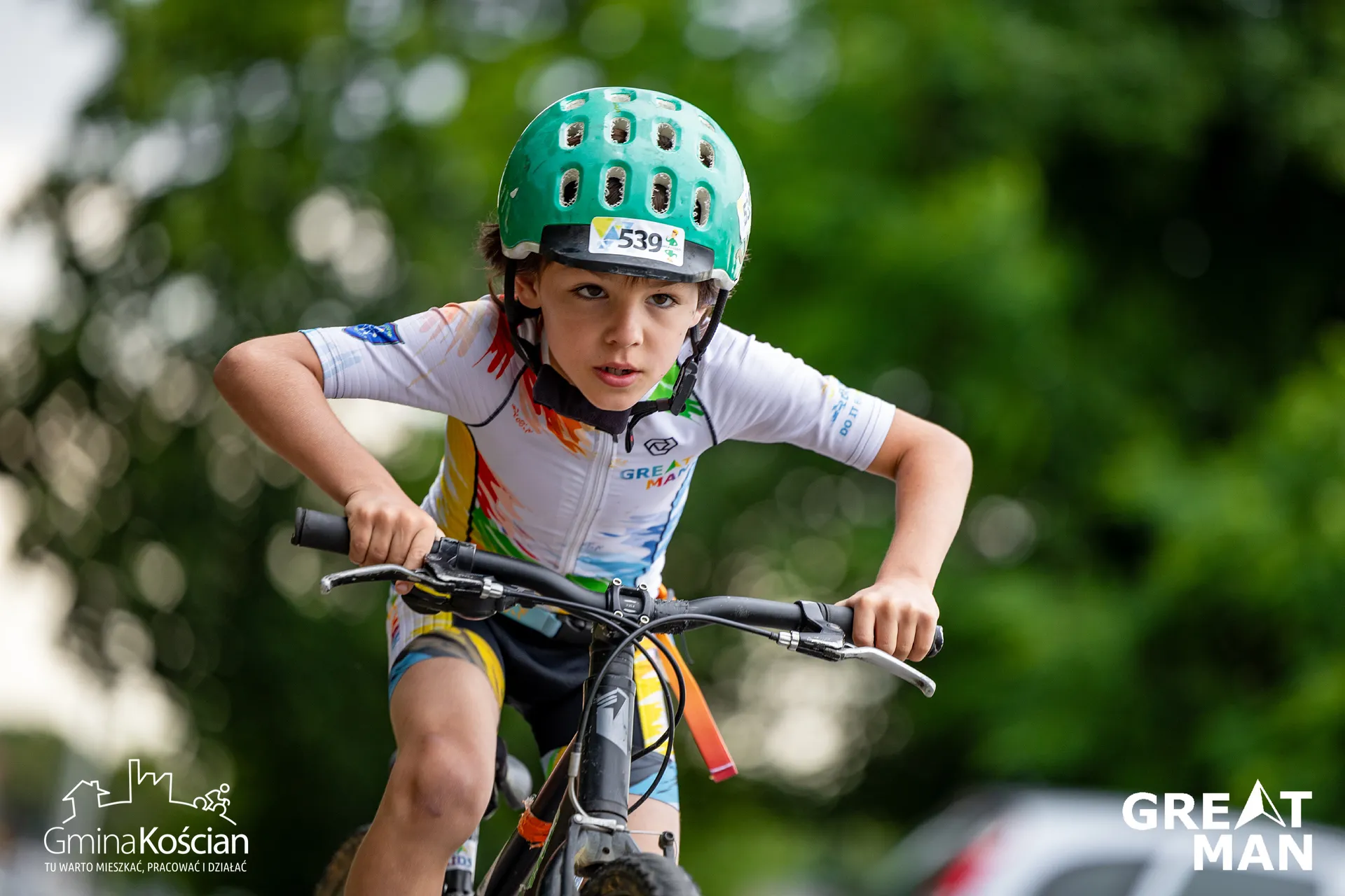 The image shows a child wearing a green helmet while riding a bike. The child is dressed in a white cycling outfit with multicolored designs. There is text and logos on the child's jersey and helmet, as well as the words “Gmina Kościan” and “GREAT MAN” visible in the image. The background is blurred, indicating motion.