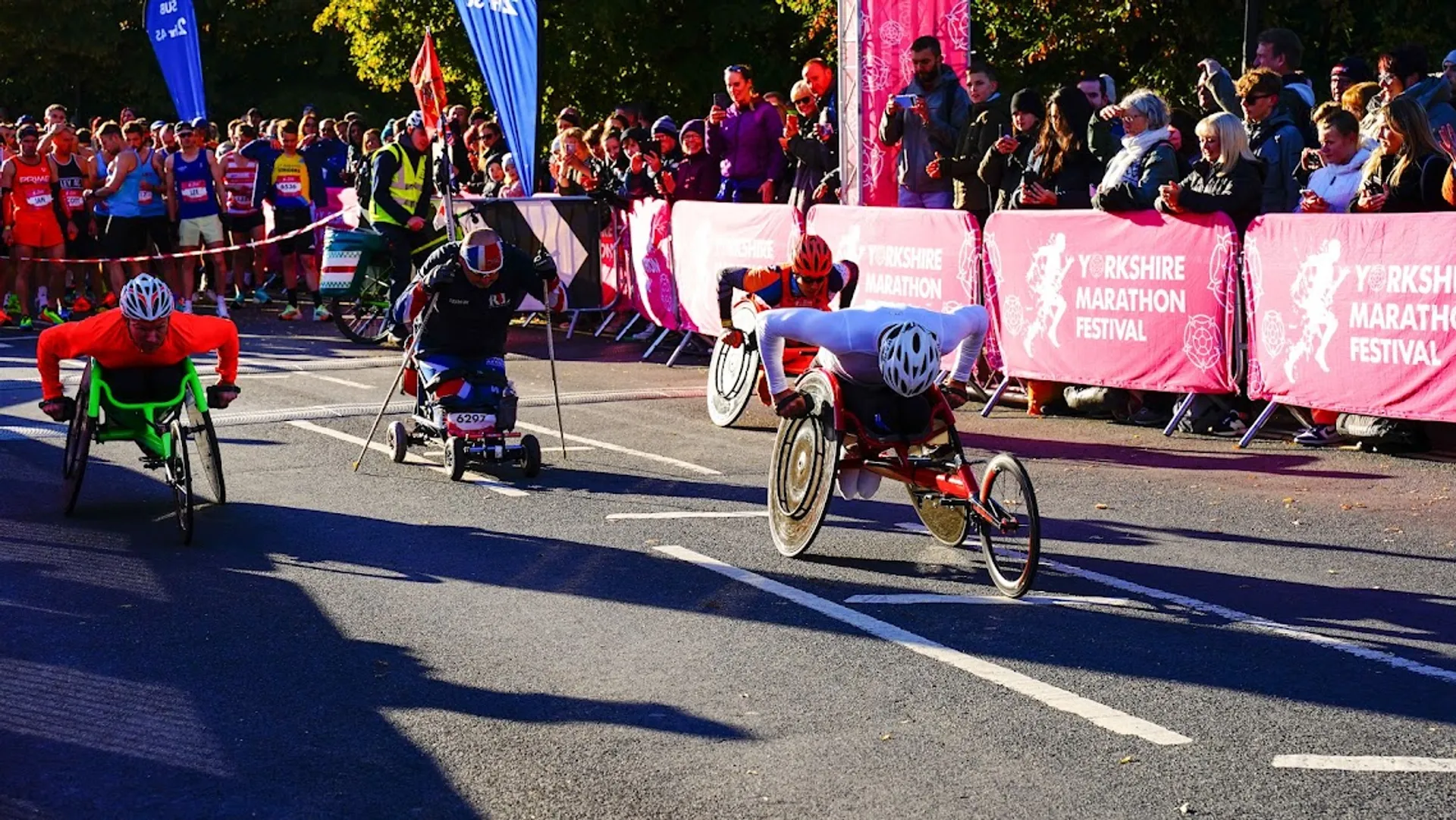 The image shows a wheelchair racing event, likely part of a marathon given the banners indicating "Yorkshire Marathon Festival." There are three athletes in racing wheelchairs at the forefront, each propelling themselves with their arms. They are on a road with a finish line in sight, marked by a banner overhead and flags along the sides. A crowd of spectators is gathered behind barriers, many of whom are watching and cheering on the racers. The scene is outdoors, in daylight, and the weather seems to be clear and sunny, contributing to the vibrant atmosphere of the event.