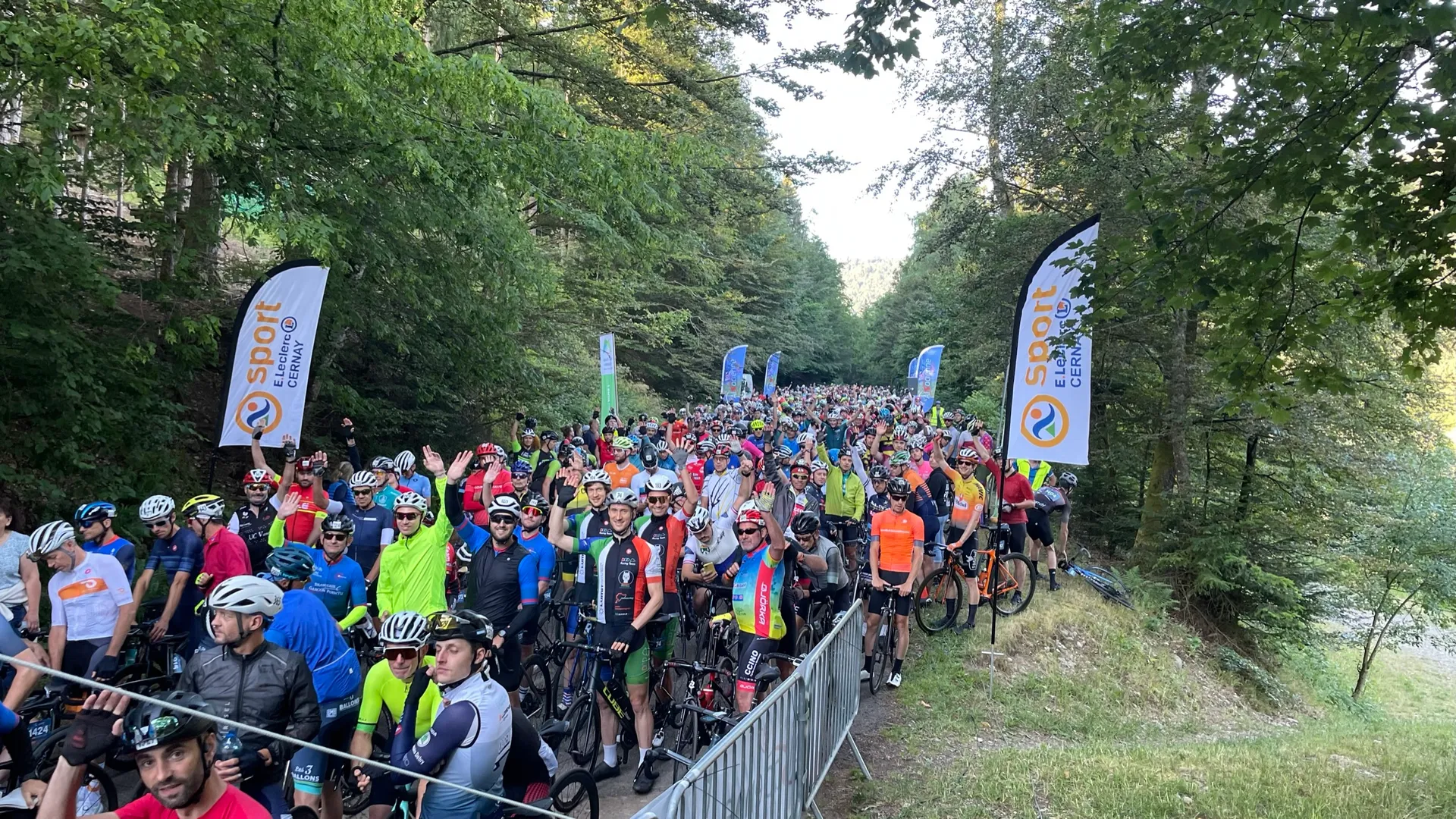 The image shows a large group of cyclists gathered at the starting line of a cycling event. They are wearing helmets and various types of cycling gear. There are flags in the background with "Sport" visible on them. The event is taking place in a wooded area.