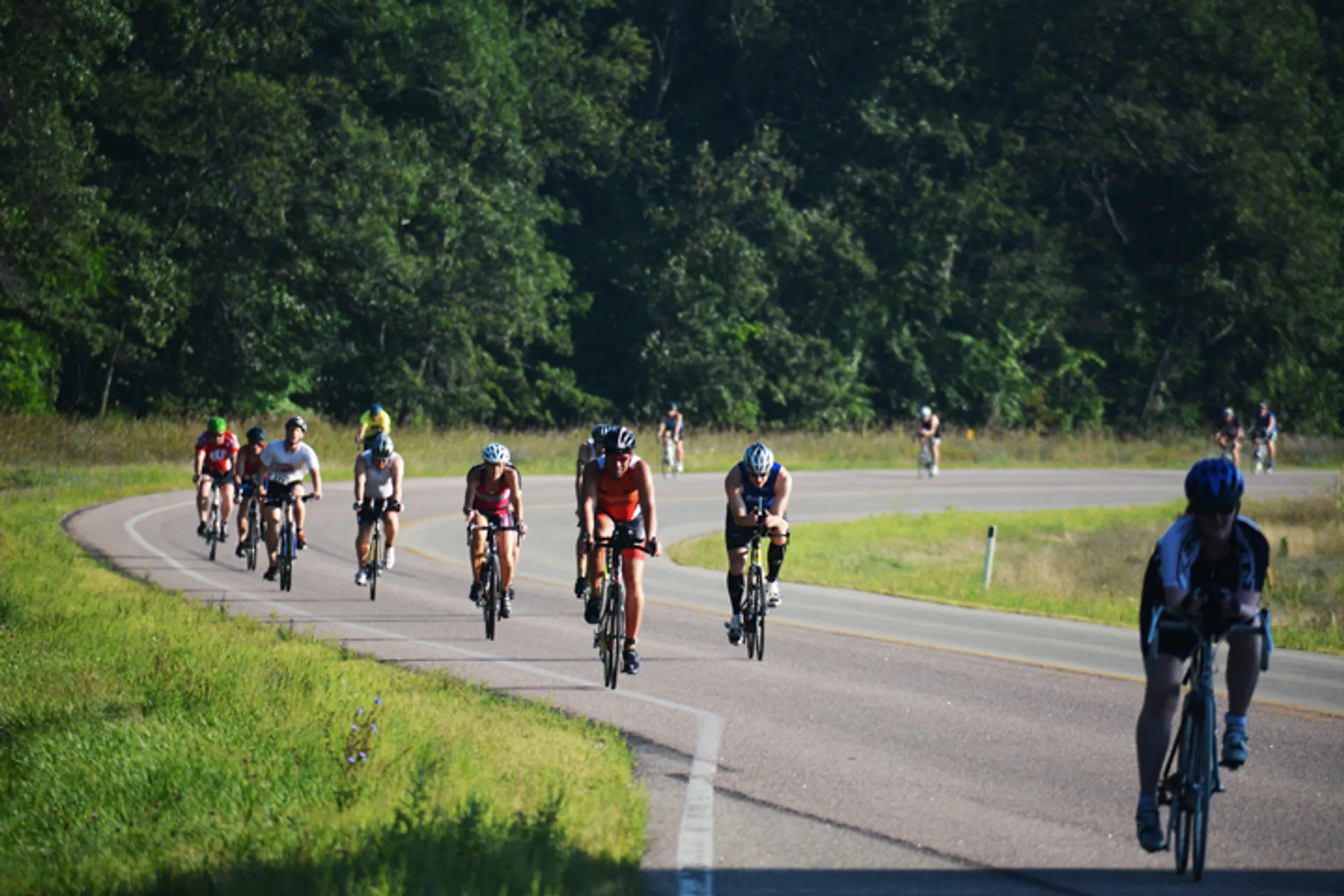 The image shows a group of cyclists riding on a road. They are spread out along the curve of the paved road, with some cyclists closer to the camera and others further away. They appear to be engaged in a cycling event or group ride, as they are wearing cycling attire and helmets, which suggests an organized activity focused on road biking. The setting is outdoors with greenery on both sides of the road, indicating a rural or park environment. It's a sunny day, and the shadows suggest it is either morning or afternoon.
