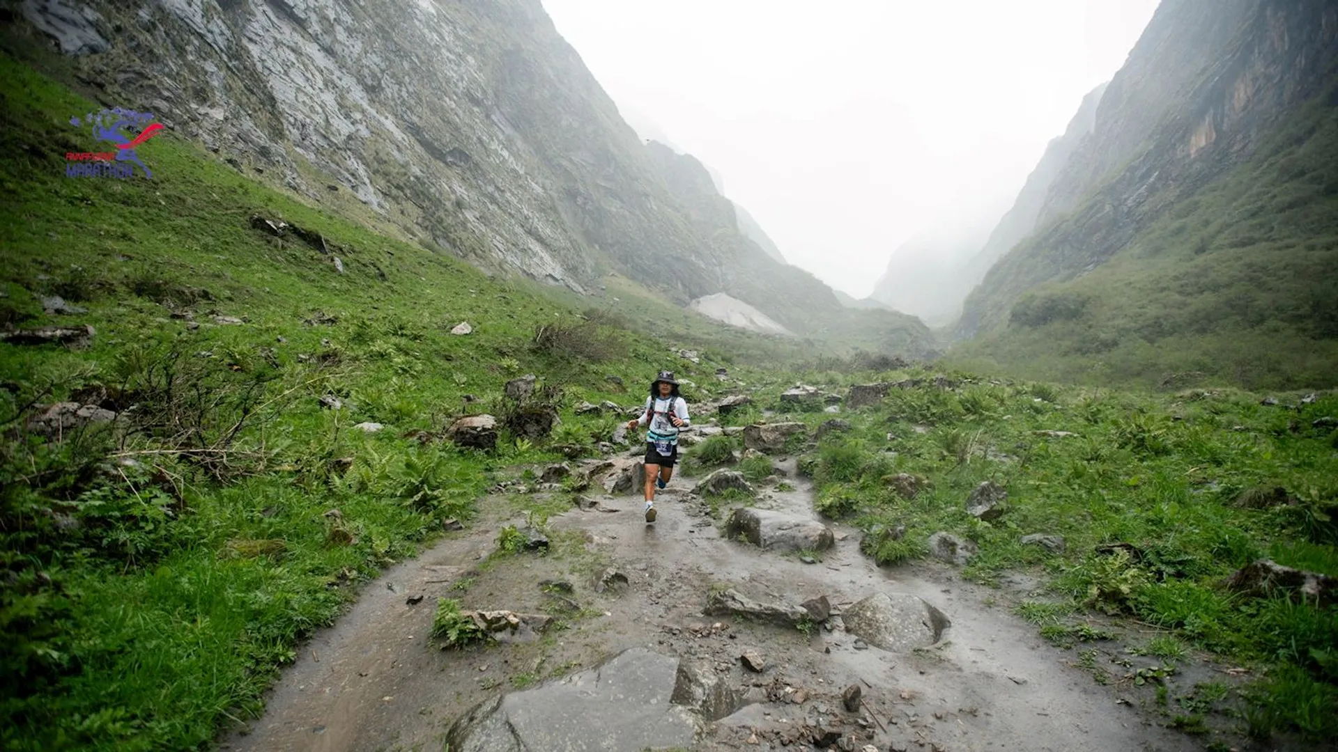 The image shows a person running on a rocky trail in a mountainous area. The surroundings are lush and green, with steep hills on either side. The weather appears to be misty or foggy, creating a serene landscape.
