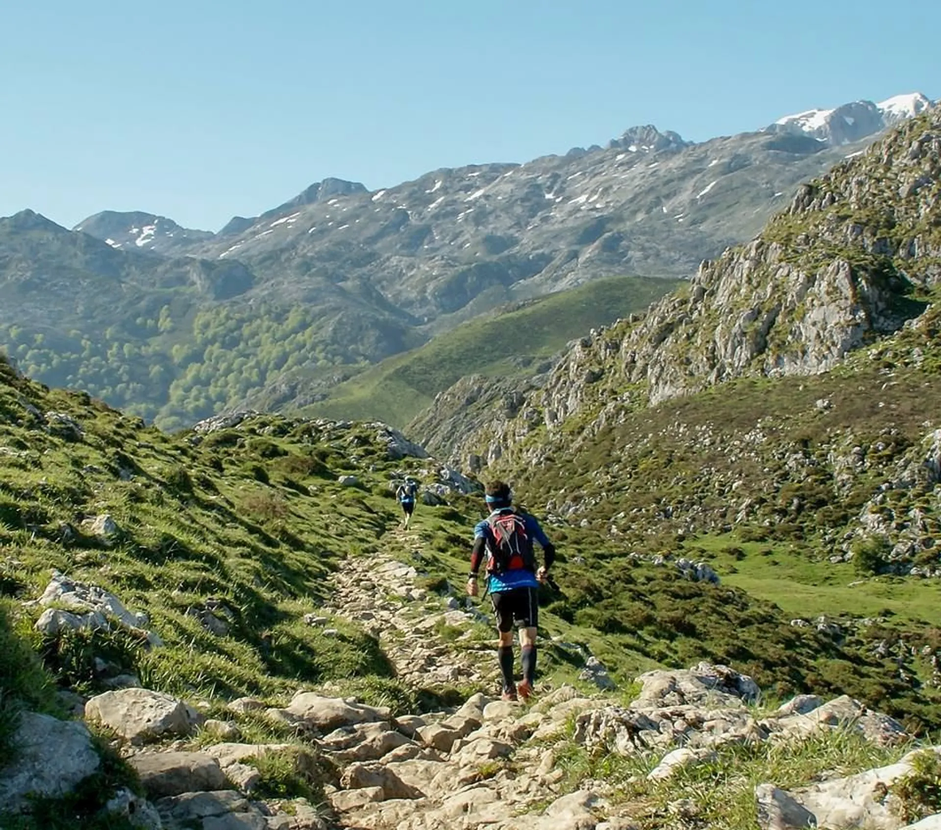 The image shows a person hiking on a rocky path in a mountainous area. The mountains in the background have a mix of rocky terrain and patches of snow. There's lush green vegetation, indicating it's likely spring or summer. The weather appears clear and sunny.