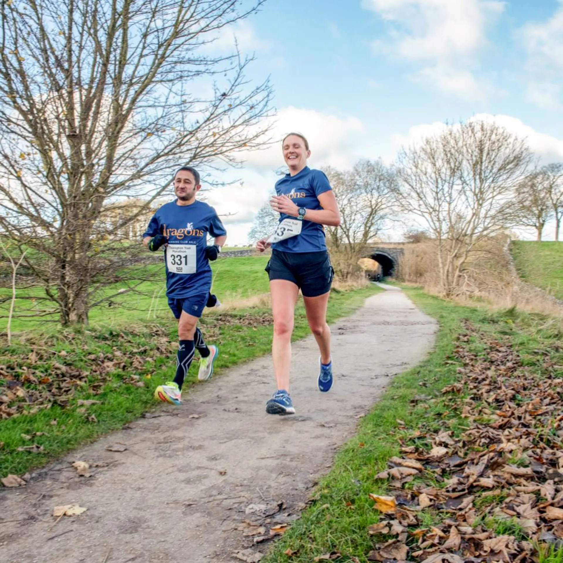 The image depicts two individuals engaged in running, likely participating in a race or a recreational run. They're on a path surrounded by grass and trees, indicating a park or rural environment. One is wearing a shirt labeled with "Dragons" which might indicate they are part of a team or club. Both have bib numbers attached to their shirts, which is common in organized running events. The weather seems to be fair with some clouds in the sky, and they appear to be in good spirits, possibly enjoying the exercise and outdoor setting. The path has leaves scattered around, suggesting it might be autumn.