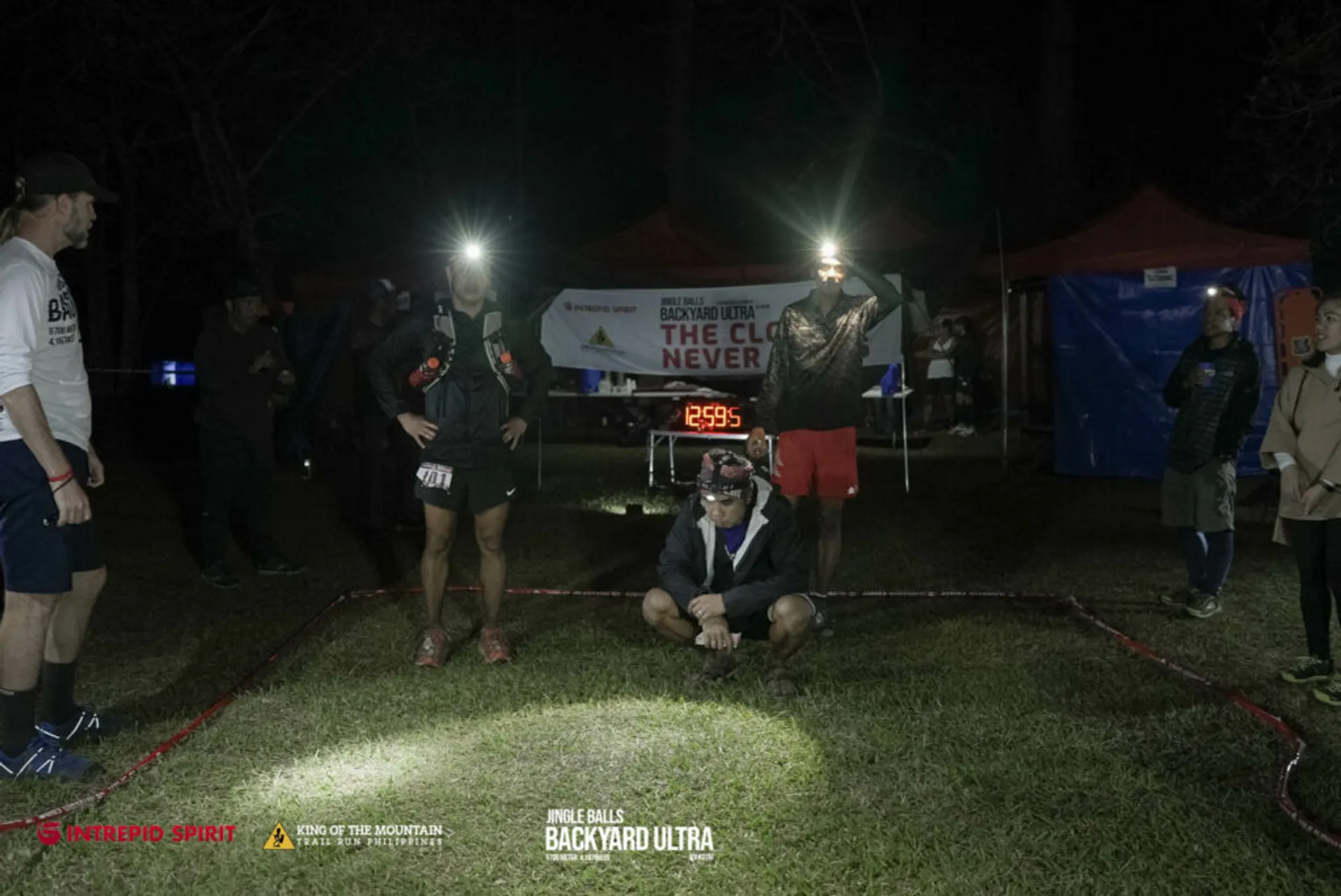 The image shows an outdoor nighttime event. There is a group of people standing and one person is crouching or kneeling at what appears to be the start or finish line of a race. The person in front is wearing a headlamp, which is common in night runs or endurance events, suggesting they might be a participant. Behind them is a banner with the text "Big's Backyard Ultra," indicating the name of the event, which is a type of ultra-endurance race. There are also other texts such as "The Closer You Get The Harder It Gets." and "Lone Pine." These are probably mottos or themes related to the race.

The atmosphere looks casual but attentive, as if the participants and attendants are