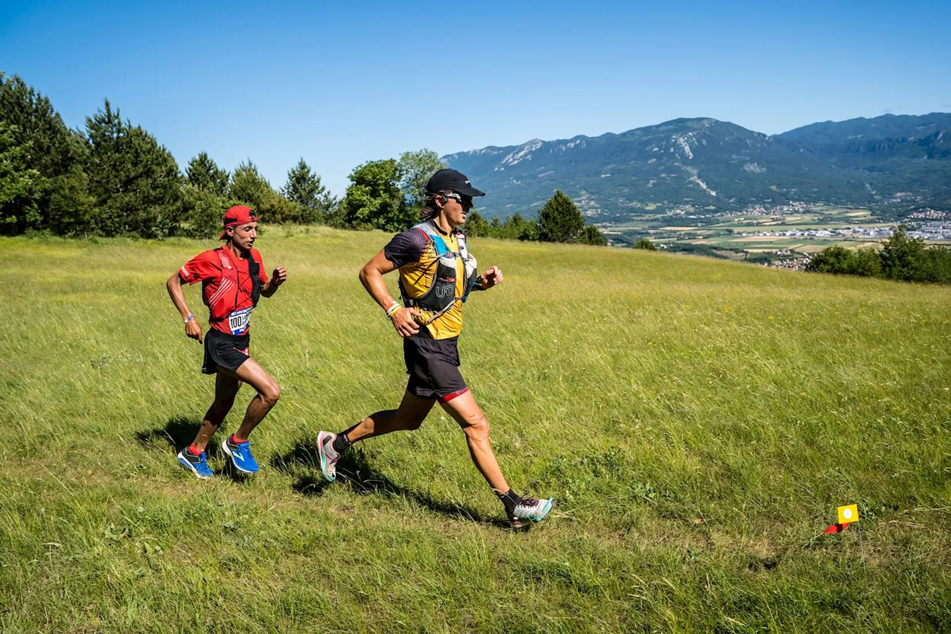 The image shows two individuals engaged in trail running, which is a sport that involves running and hiking over trails. They are both wearing running attire suitable for the activity, such as shorts, t-shirts, running shoes, and one of them has a cap and sunglasses to protect from the sun. The runner in the foreground also appears to be carrying a hydration pack on his back, which is common for long-distance trail running to stay hydrated. They are running through a grassy field with a marked running route indicated by small flags or markers on the ground. In the background, there's a panoramic view of a mountain range under a clear blue sky, suggesting that the location offers a scenic environment for the sport.