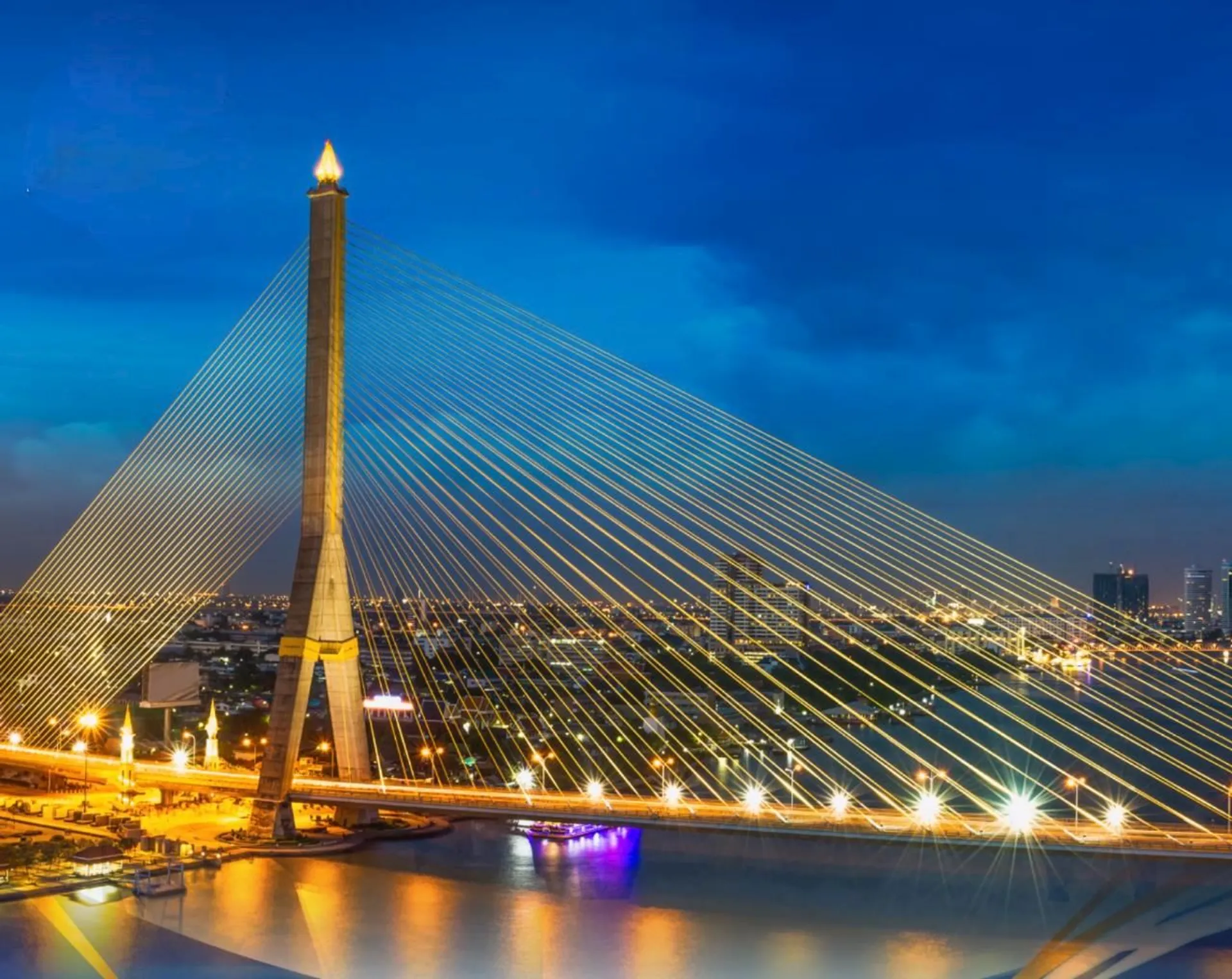 The image depicts a brightly lit cable-stayed bridge at night. The bridge features a prominent tall tower with numerous cables fanning out to the deck. It spans a body of water, and city lights are visible in the background, creating a vibrant nighttime cityscape.
