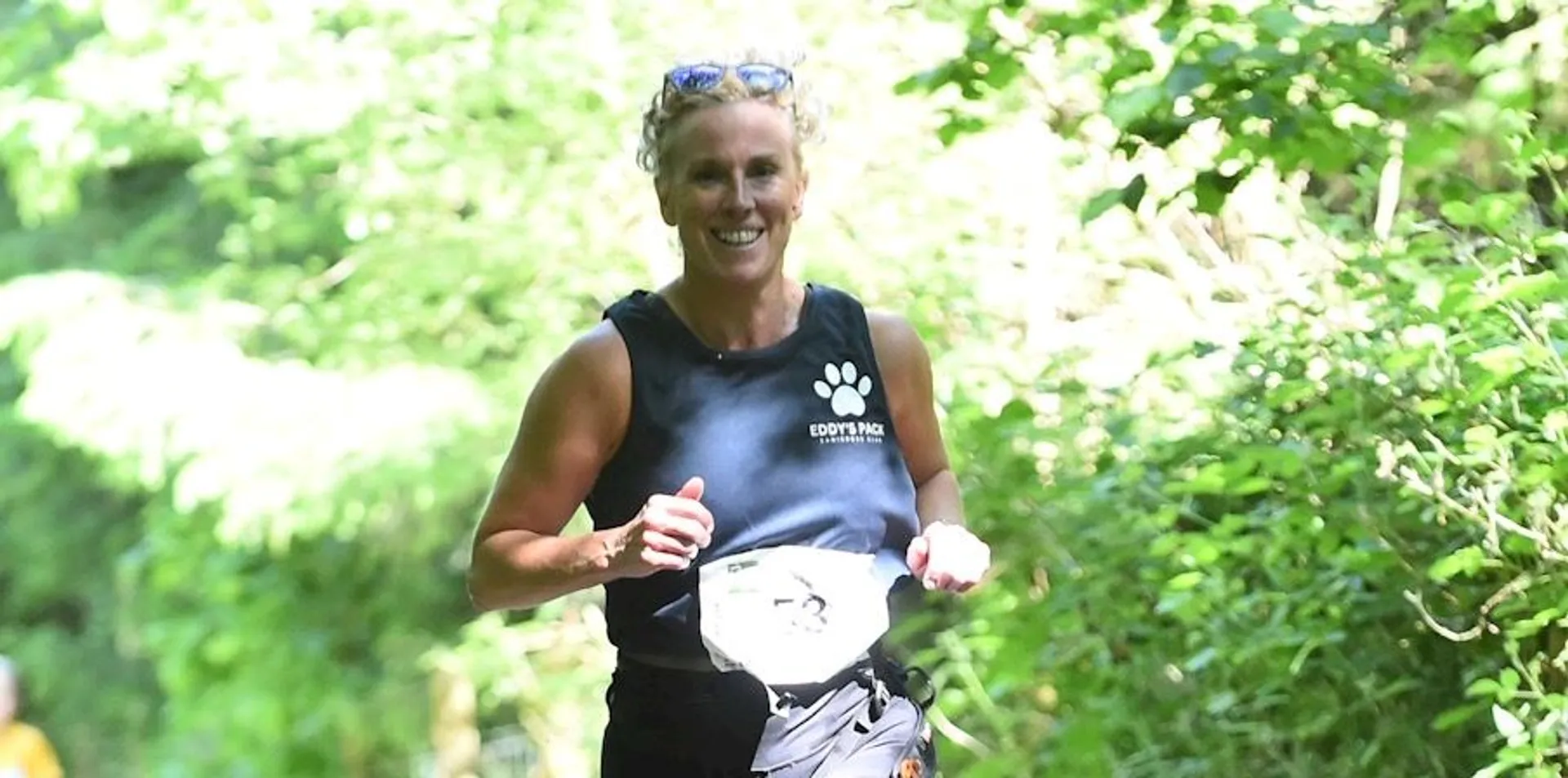 The image shows a person running outdoors along a path surrounded by greenery. They are wearing a sleeveless top with a paw print logo and appear to have a race bib on their shorts. The person looks happy and is holding something in their hand.