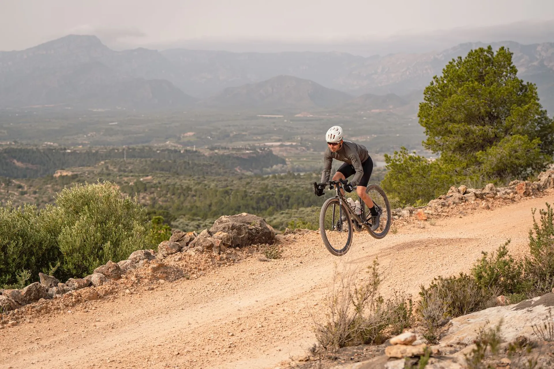 The image features a person riding a mountain bike on a dirt trail. The rider is wearing a helmet for safety and sports attire suitable for biking. The background reveals a scenic view with mountains in the distance and partially cloudy skies. The terrain appears dry and rocky, characteristic of mountainous or hilly landscapes that are popular for mountain biking.