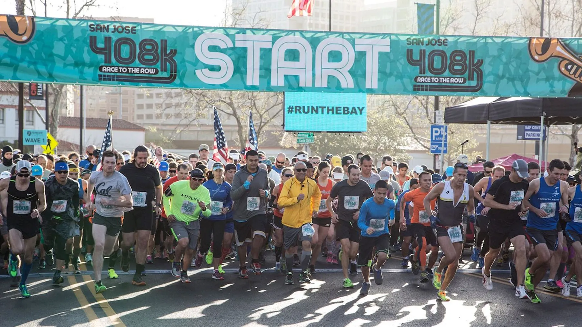 The image shows a group of runners at the starting line of a race titled "San Jose 408k Race to the Row." A banner overhead reads "START," and there is a hashtag "#RUNTHEBAY" displayed. The runners are in a street setting, prepared to begin the race.