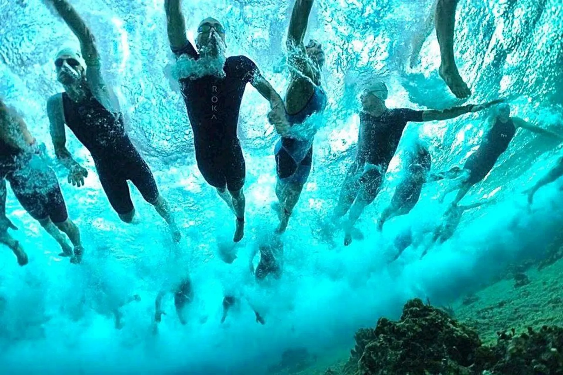 The image depicts a group of swimmers from an underwater perspective. They are all wearing swimwear, possibly wetsuits, and are in the midst of what looks like an open water swimming event or a group swim. The water is a clear turquoise color, and we can see bubbles and splashes around the swimmers, indicating their active movement. The sunlight is filtering through the water from above, creating an interesting pattern of light and shadow. The environment suggests a natural water body rather than a swimming pool.