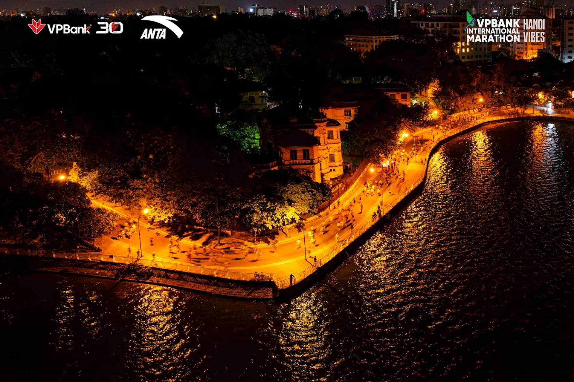 The image shows a nighttime scene of a curving walkway alongside a body of water. It appears to be in an urban area, with the presence of lights and some buildings surrounded by trees on one side. Streetlights line the walkway, casting an orange glow and reflecting on the water's surface. The scene is peaceful and scenic, with the lights creating a serene atmosphere. There's also text at the top of the image that mentions "VPBank Hanoi International Marathon VIBES," indicating that this image could be linked to a specific event, likely a marathon in Hanoi.