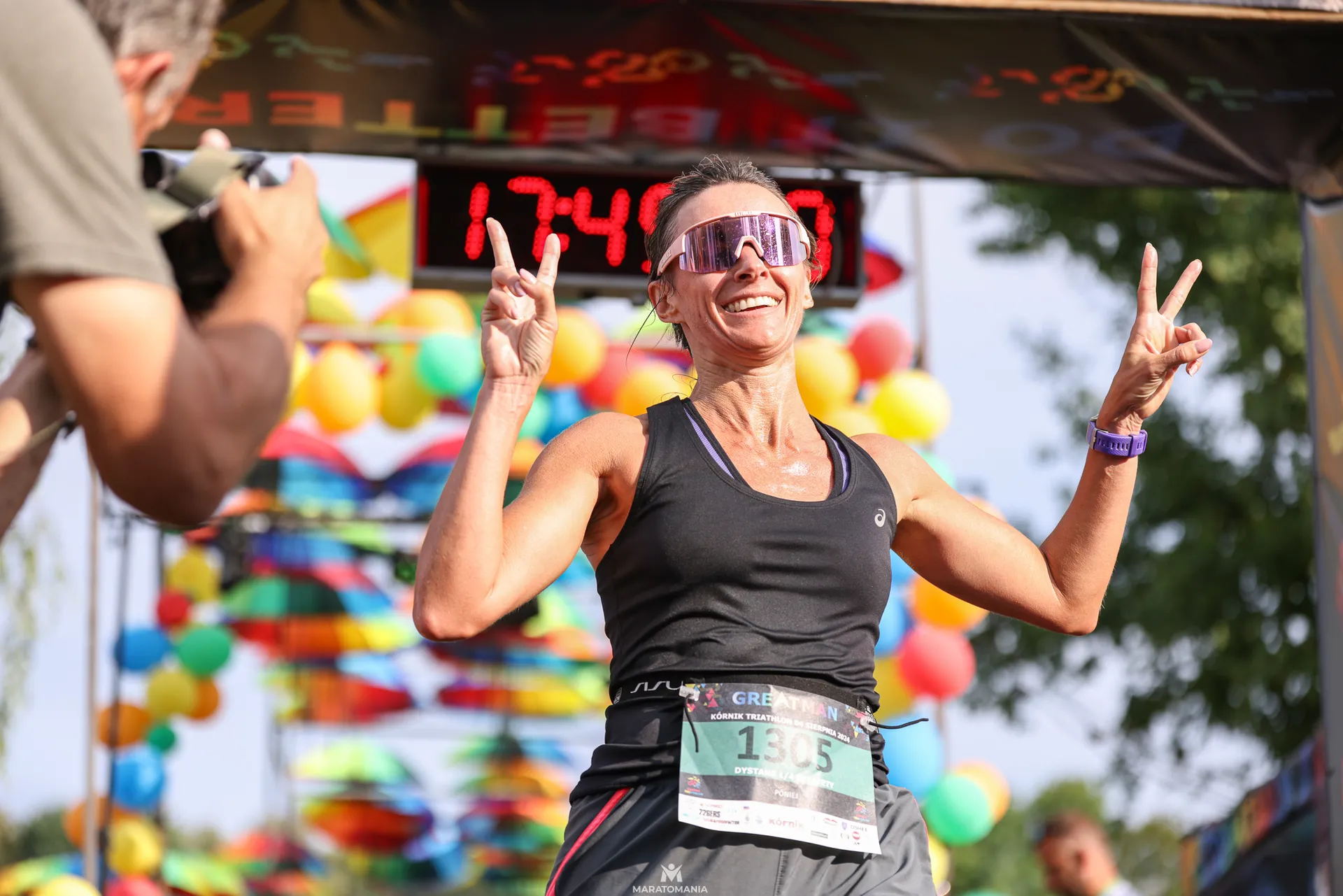 The image shows a person finishing a race. They are smiling, wearing sporty sunglasses, and making peace signs with both hands. The background has colorful balloons and a timer shows "1:47:42". There are other people visible, including someone with a camera.

