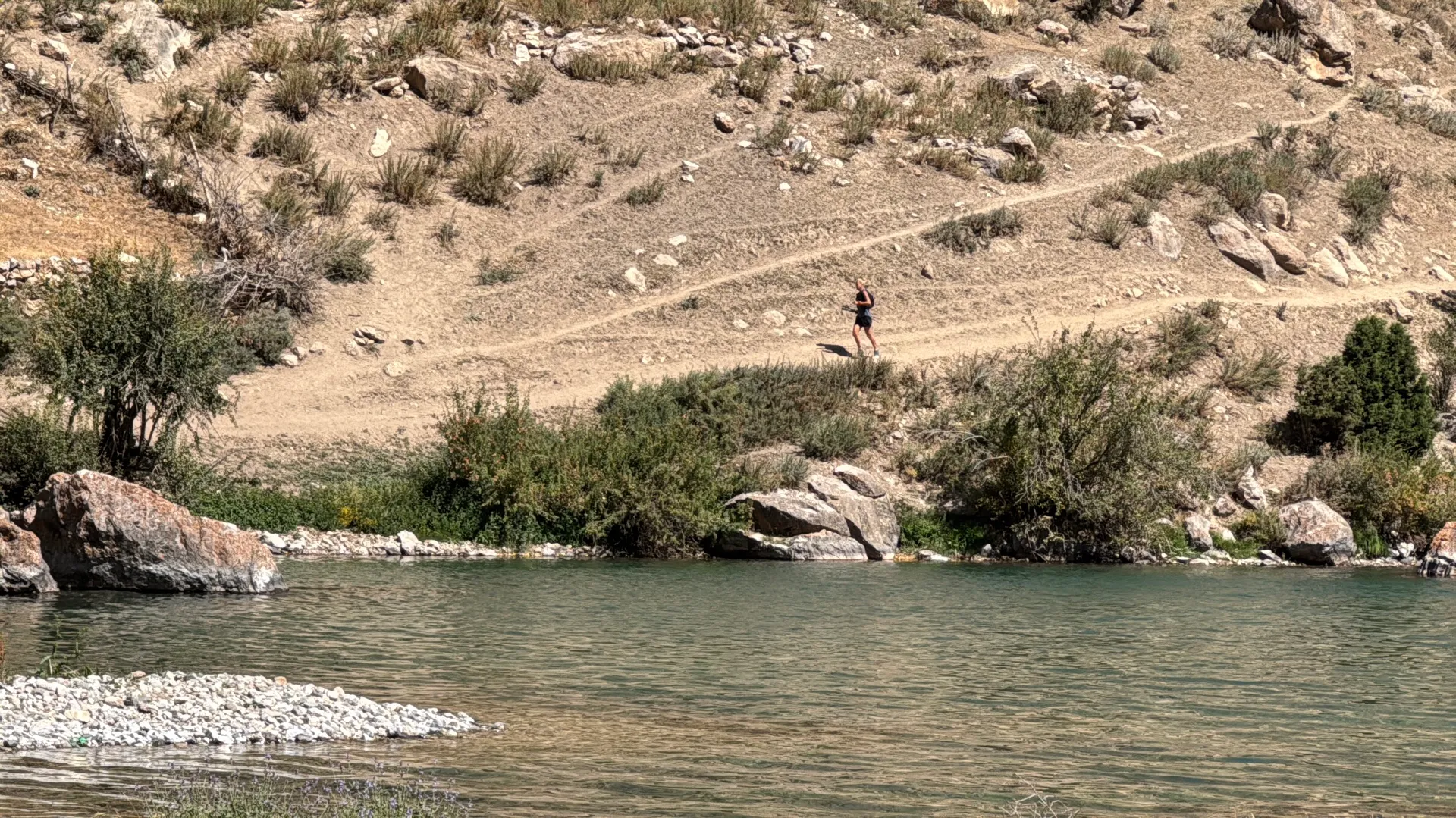 The image shows a person standing on a dirt path near a rocky hillside. In the foreground, there's a body of water, possibly a river or lake, with rocky banks. Vegetation is sparse, indicating a dry, possibly arid environment.