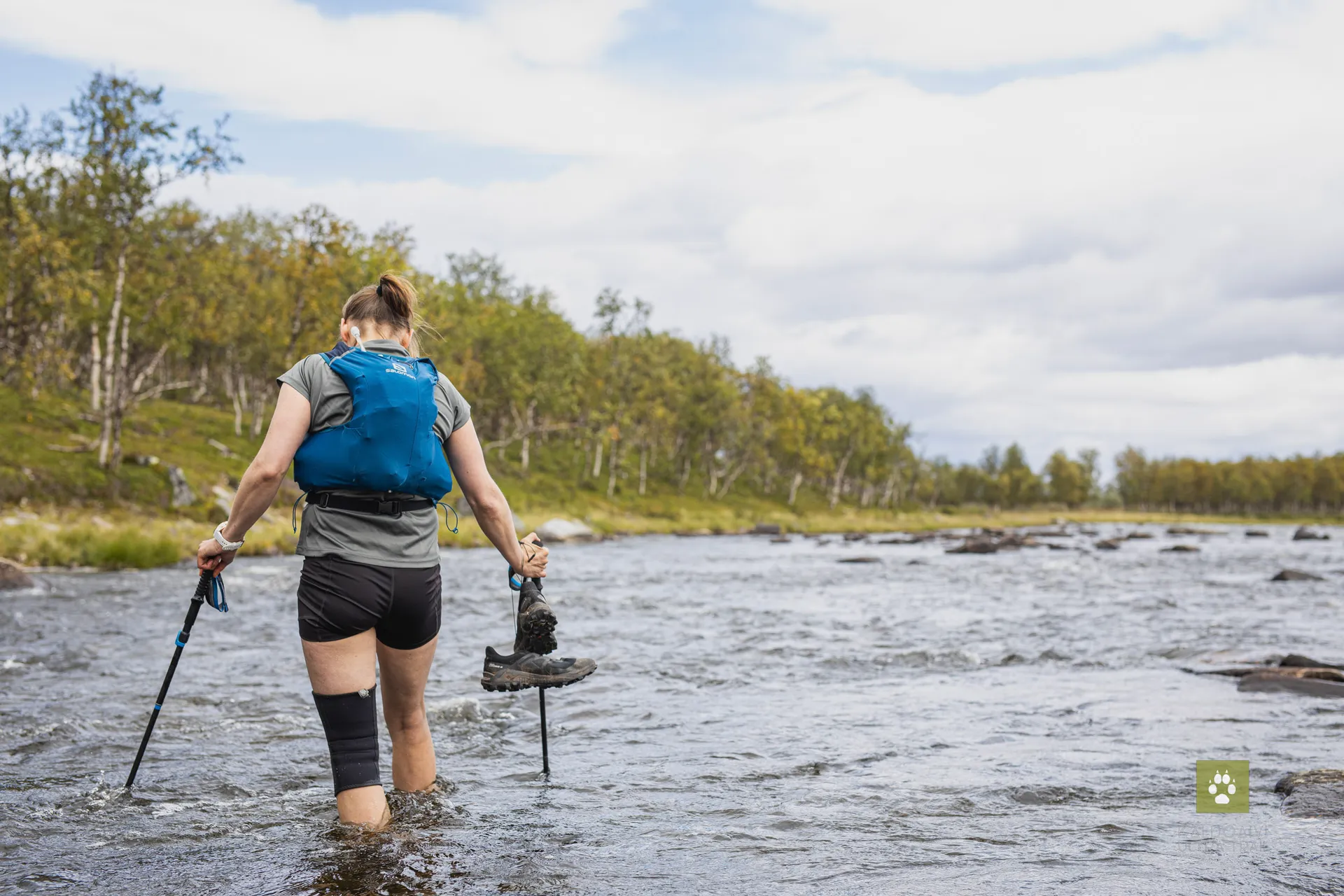The image depicts a person from behind who is wading through a shallow river. The person appears to be equipped for hiking or trekking, wearing shorts and a t-shirt, and carrying a blue backpack with a visible hydration pack hose. They are using hiking poles for stability, and it seems they have lifted one of their feet, possibly to take a step or to avoid getting their shoe wet. The river is set in a natural environment with trees and foliage in the background, and the sky is partly cloudy. The scene suggests a sense of adventure and outdoor exploration.