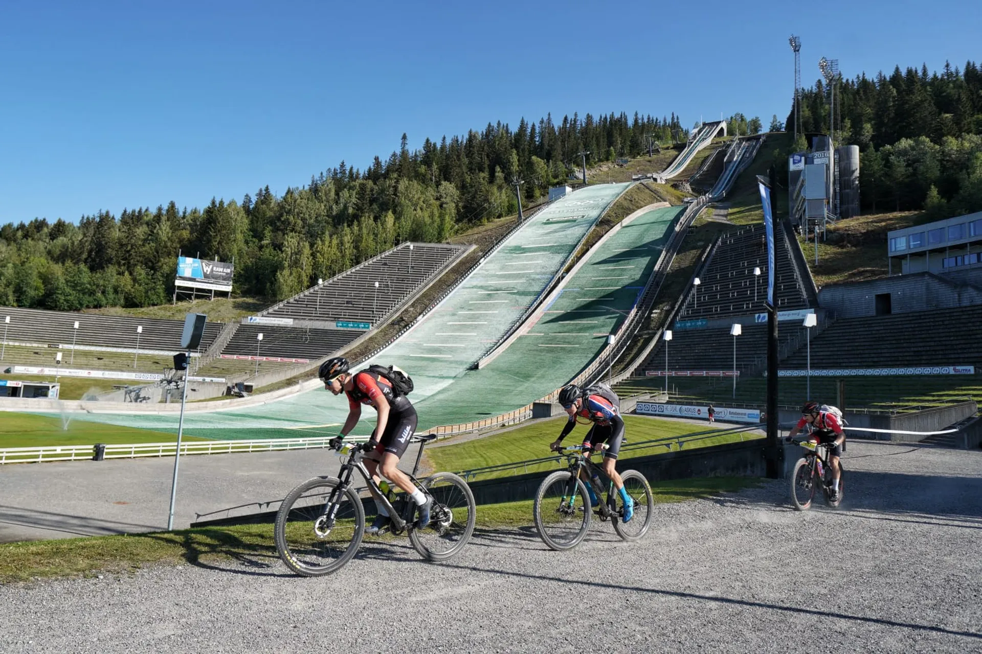 This image features two cyclists riding on a road in front of a large ski jump