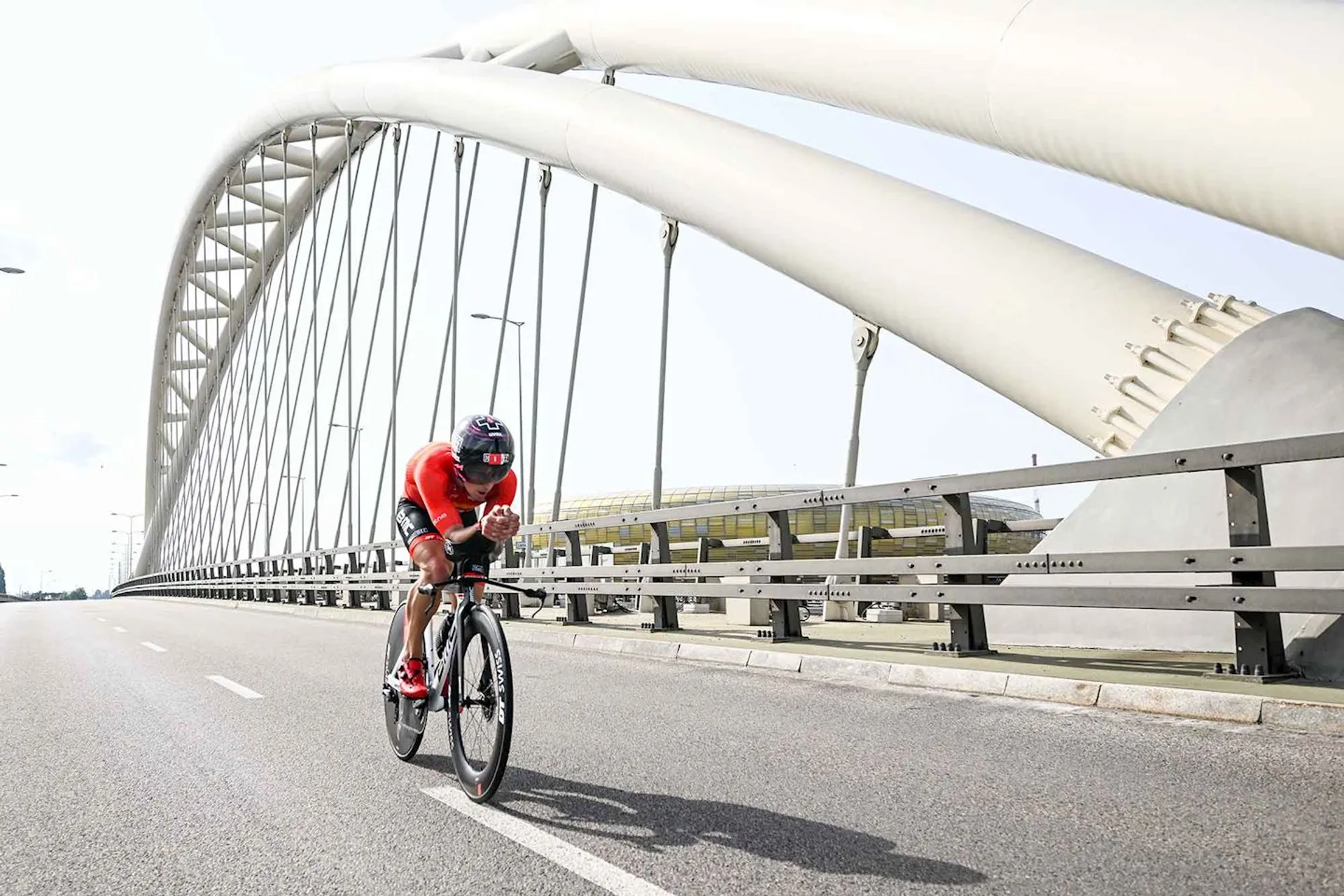 The image shows a cyclist in a red outfit riding a road bike on a bridge. The bridge has a large, modern arch structure. The background features a clear sky.