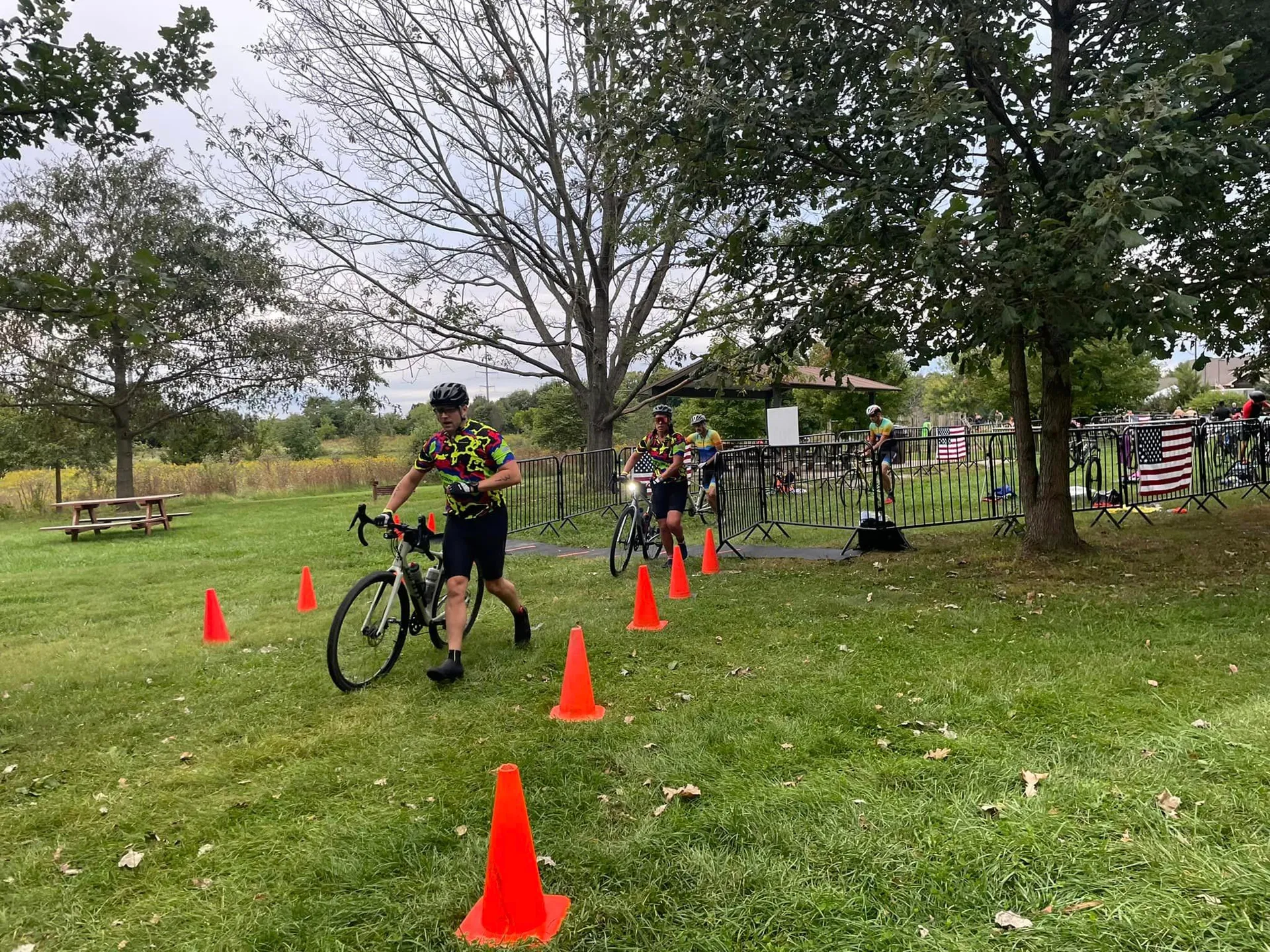 The image shows a group of cyclists at what appears to be a biking event or race. The cyclists are wearing matching cycling outfits with bright and colorful patterns. One cyclist is in the foreground, dismounting or maneuvering around orange traffic cones on a grassy area, which suggests this could be part of a cyclocross race or a skills section of a cycling event. Trees and a cloudy sky can be seen in the background, along with what looks like a festival or event setup, including tents and other structures that are often associated with outdoor events. There appear to be spectators or other participants in the background, indicating a social or competitive gathering.