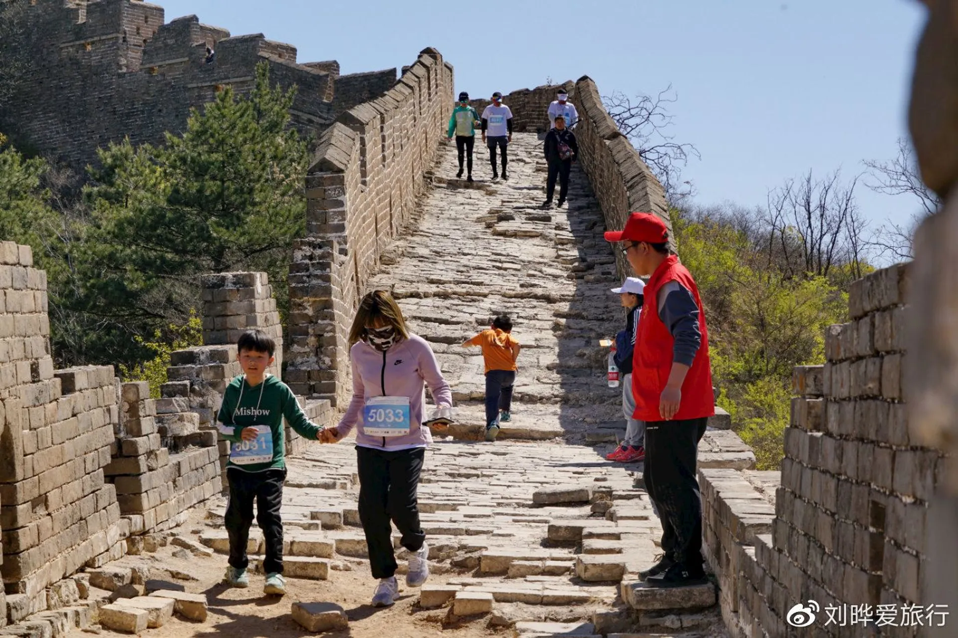 In the image, you can see people who appear to be visitors or tourists walking on what looks like the Great Wall of China, given the distinctive architecture of the wall. Some individuals are walking upwards along the sloped pathway of the wall, which appears to be made of stone and bricks. The wall follows the natural contours of the landscape, which is hilly and covered with some greenery, suggesting it could be spring or summer. The sky is blue, indicating a clear day.

There is a mix of adults and children in the picture. A woman in a red jacket appears to be standing and observing or waiting, while in the foreground two children are holding hands walking down the slope. One of them is wearing a bib with a number, suggesting