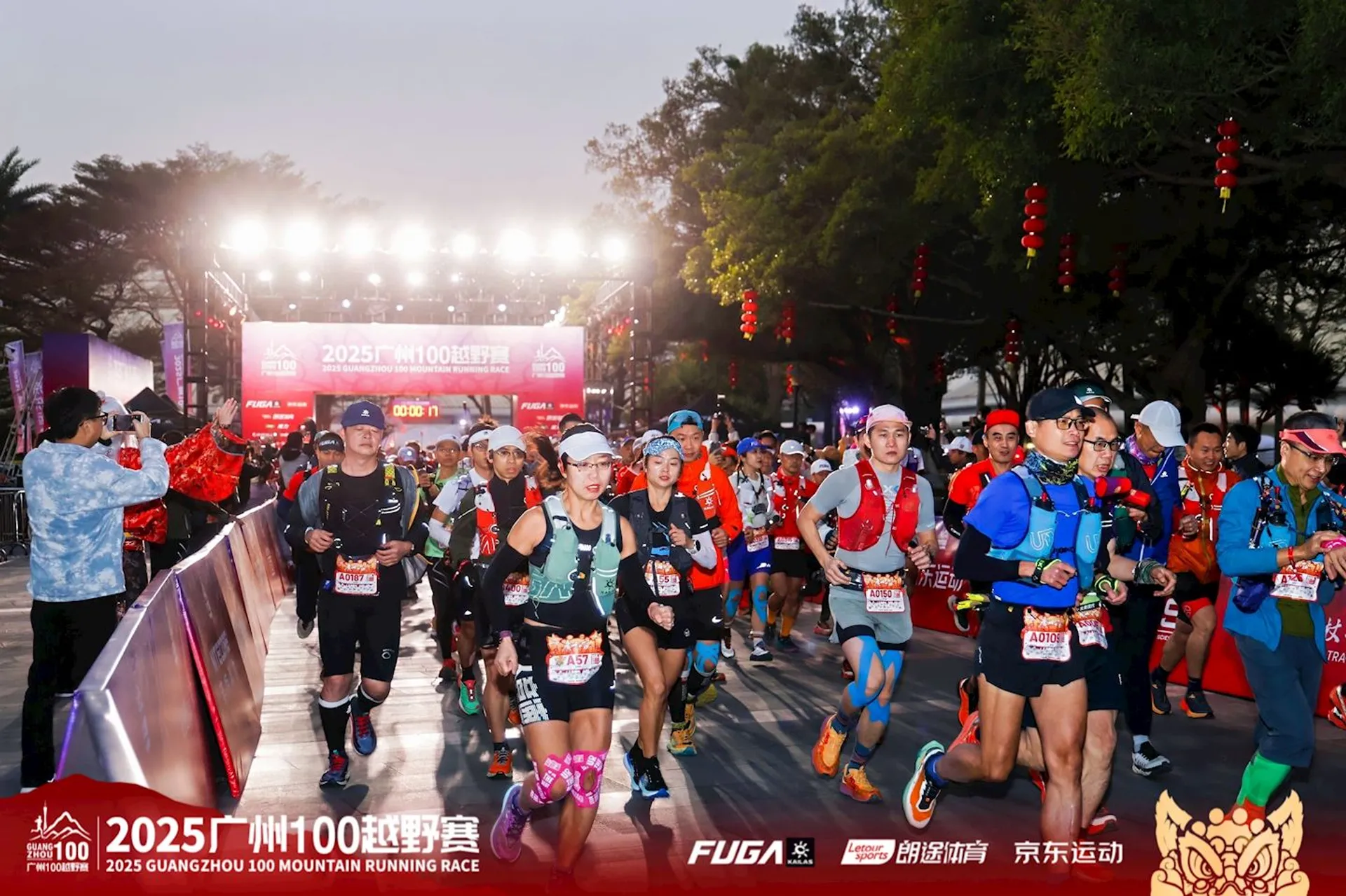 The image shows a group of runners participating in the 2025 Guangzhou 100 Mountain Running Race. They are at the starting line, dressed in athletic gear, with some wearing hydration packs and headlamps. The scene is vibrant, with lights and decorations, likely indicating an early morning or evening start. Banners and sponsor logos are visible in the background.