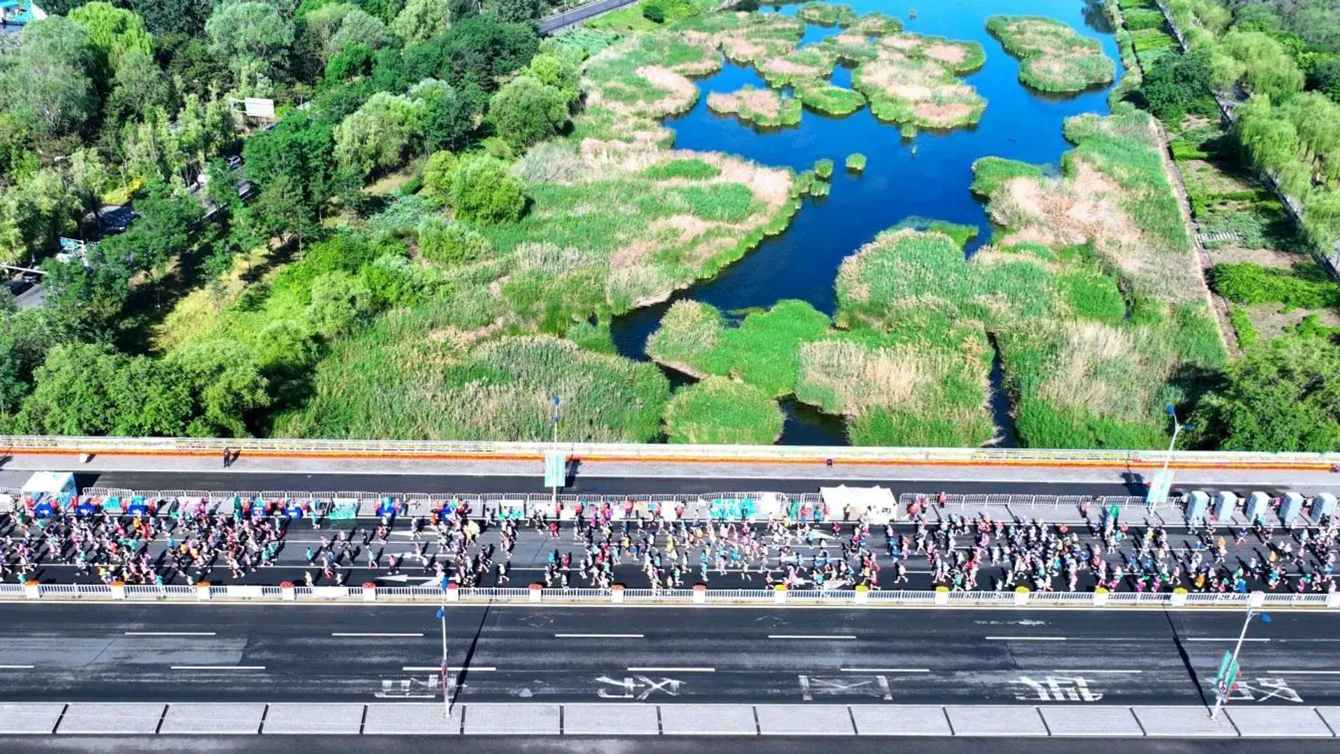 The image shows an aerial view of a road with numerous runners participating in what appears to be a marathon or a race. The road is beside a body of water with green vegetation and small patches of land. The landscape is lush with trees and greenery surrounding the area.