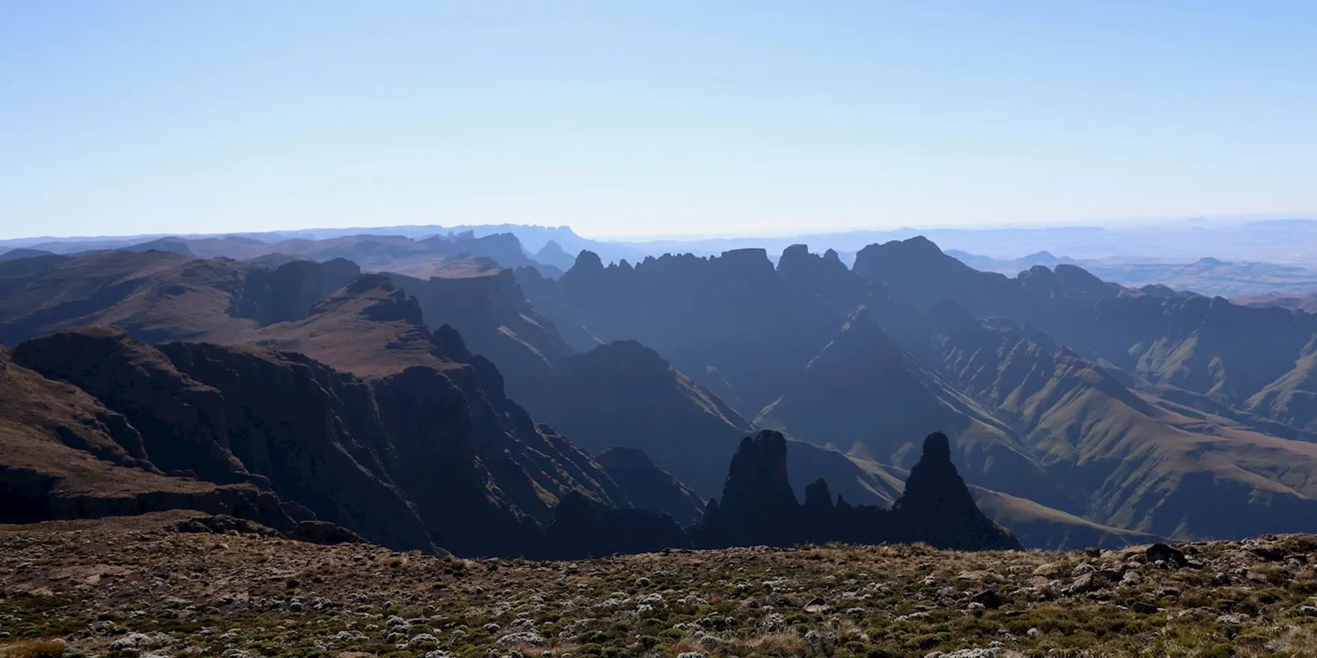 This image depicts a rugged mountain landscape with dramatic peaks and valleys. The lighting suggests it might be early morning or late afternoon, casting shadows across the mountainous terrain. The foreground shows rocky terrain, while the distant background fades into a hazy horizon.