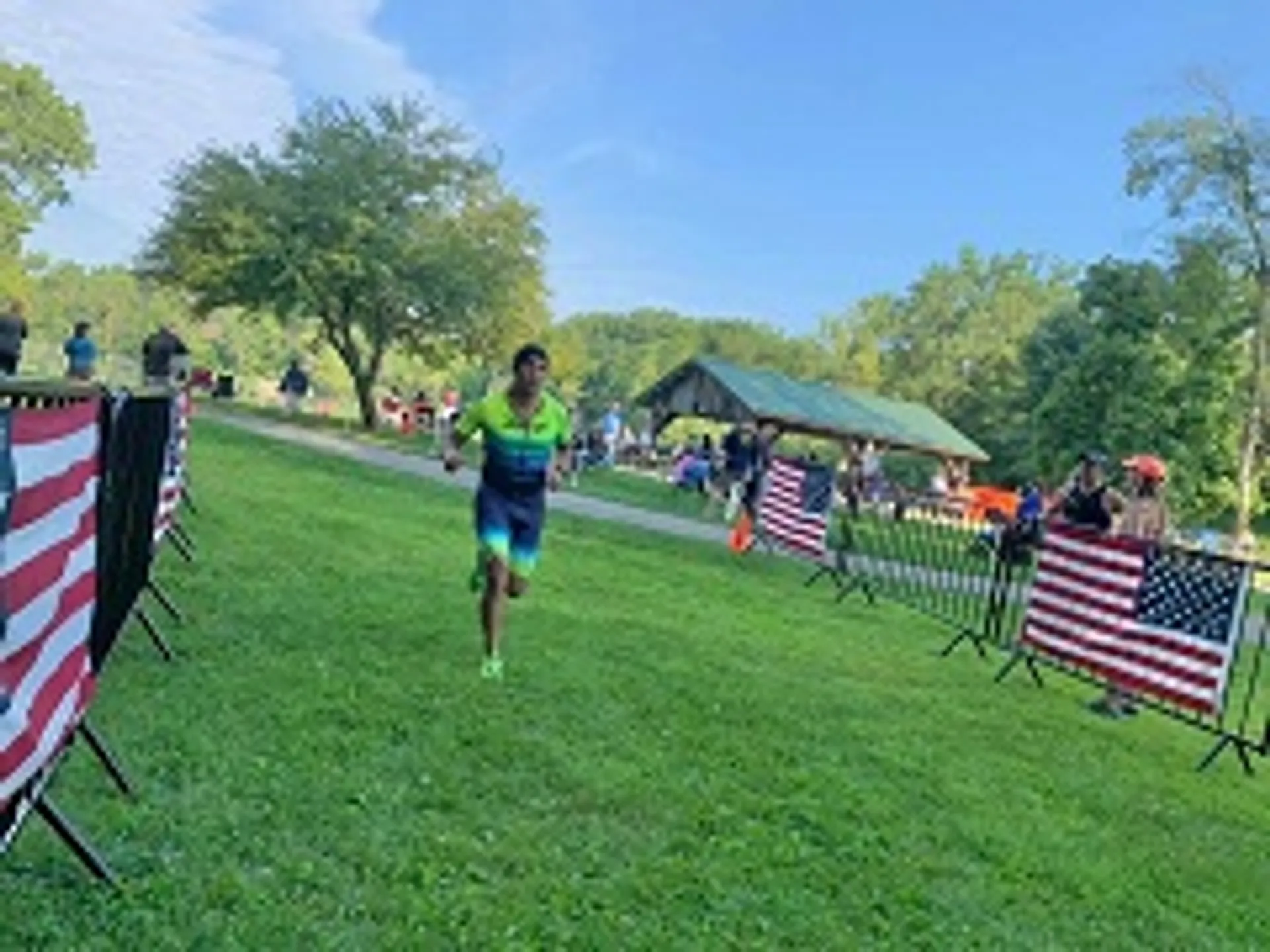 The image shows an outdoor setting, possibly a park, with a person running or jogging on the grass. The runner is wearing athletic clothing, indicating some form of exercise or participation in a running event. On the left side of the image, there are American flags attached to a barrier or fencing, which suggests the event may be related to a patriotic holiday or celebration in the United States. In the background, there are various people gathered around a shelter or pavilion, some trees, and open green space. The setting appears to be informal and likely community-oriented.