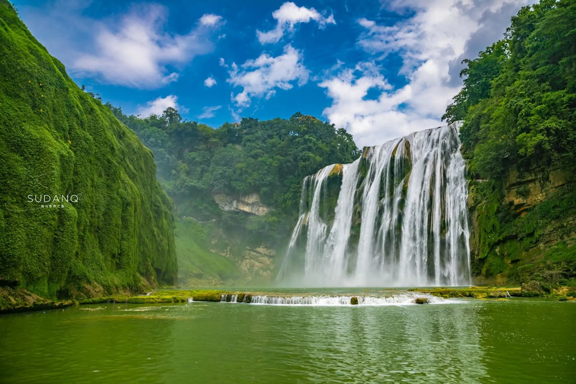The image shows a scenic waterfall cascading down a lush, green cliff into a body of water. The area is surrounded by dense vegetation, and the sky is bright with some clouds. The atmosphere is serene and natural.