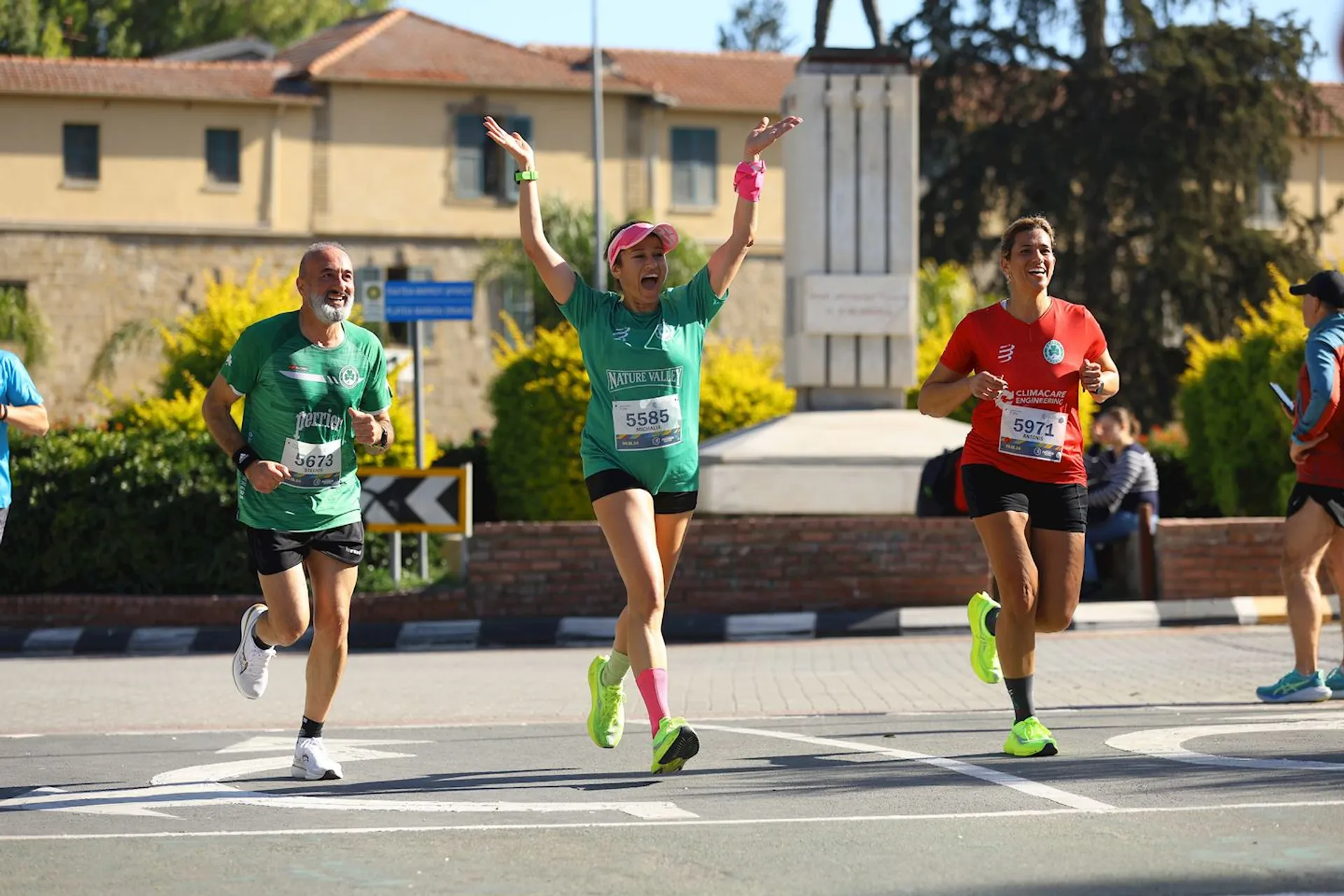 This image shows three people participating in a running event. They are wearing athletic gear and numbered bibs. The person in the center has their arms raised in excitement. There are buildings and trees in the background.