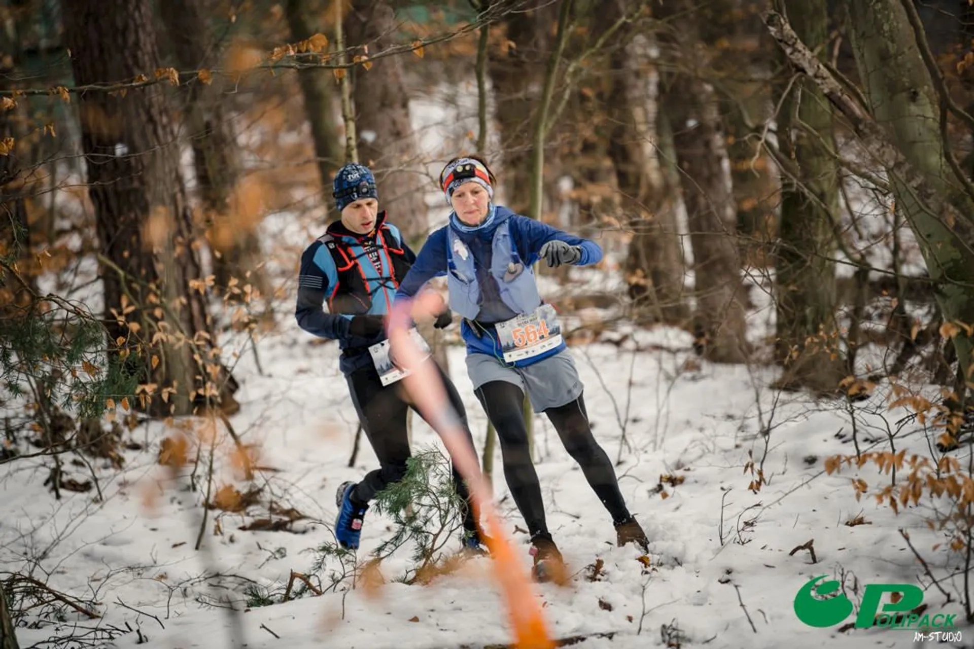 The image shows two people running through a snowy forest. They seem to be participating in a trail race or event, as indicated by the race numbers on their attire. The ground is covered in snow, and they are dressed in winter running gear. There's a blurred orange tape or line in the foreground, possibly part of the race marking.
