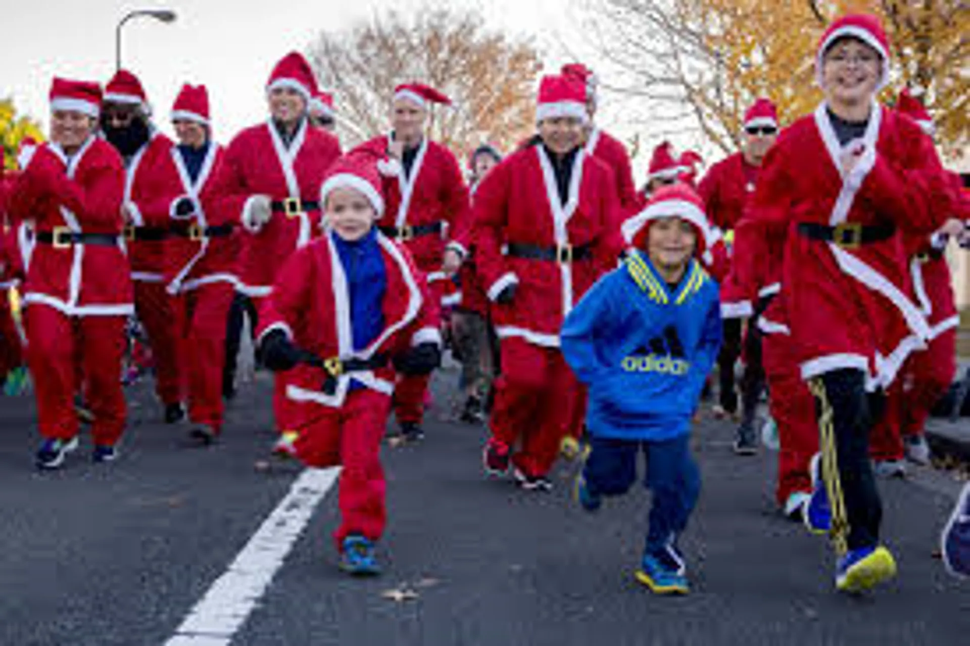 In this image, there is a group of people participating in a festive event, most likely a Santa run, where participants dress up in Santa Claus costumes and run together. The group consists of adults and children; they are all wearing Santa hats, and the majority are dressed in the traditional red and white Santa suits. Among the crowd, two children are prominent in the foreground, one dressed in a blue sports outfit and the other in a Santa costume, both running and seemingly enjoying the event. They are on a street lined with leafless trees, indicating it might be a cold season or late autumn/winter period. The atmosphere appears to be joyful and fun.