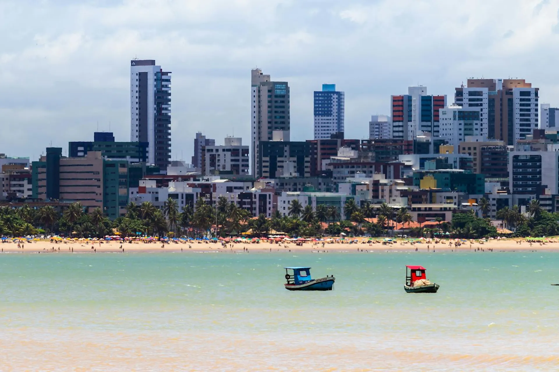 The image shows a coastal cityscape with high-rise buildings in the background and a beach in the foreground. There are two small boats with colorful sails on the water. There are also people visible along the shoreline.