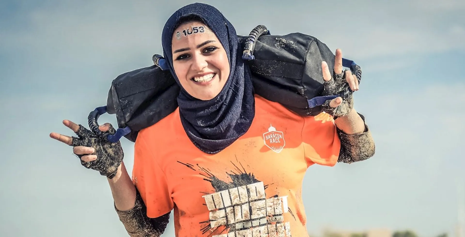 The image shows a person wearing a hijab and an orange T-shirt, carrying a sandbag over their shoulders. They appear to be participating in a challenge or race, as they are wearing a numbered race bib. Their hands and clothes are muddy, indicating involvement in an obstacle course or similar event.
