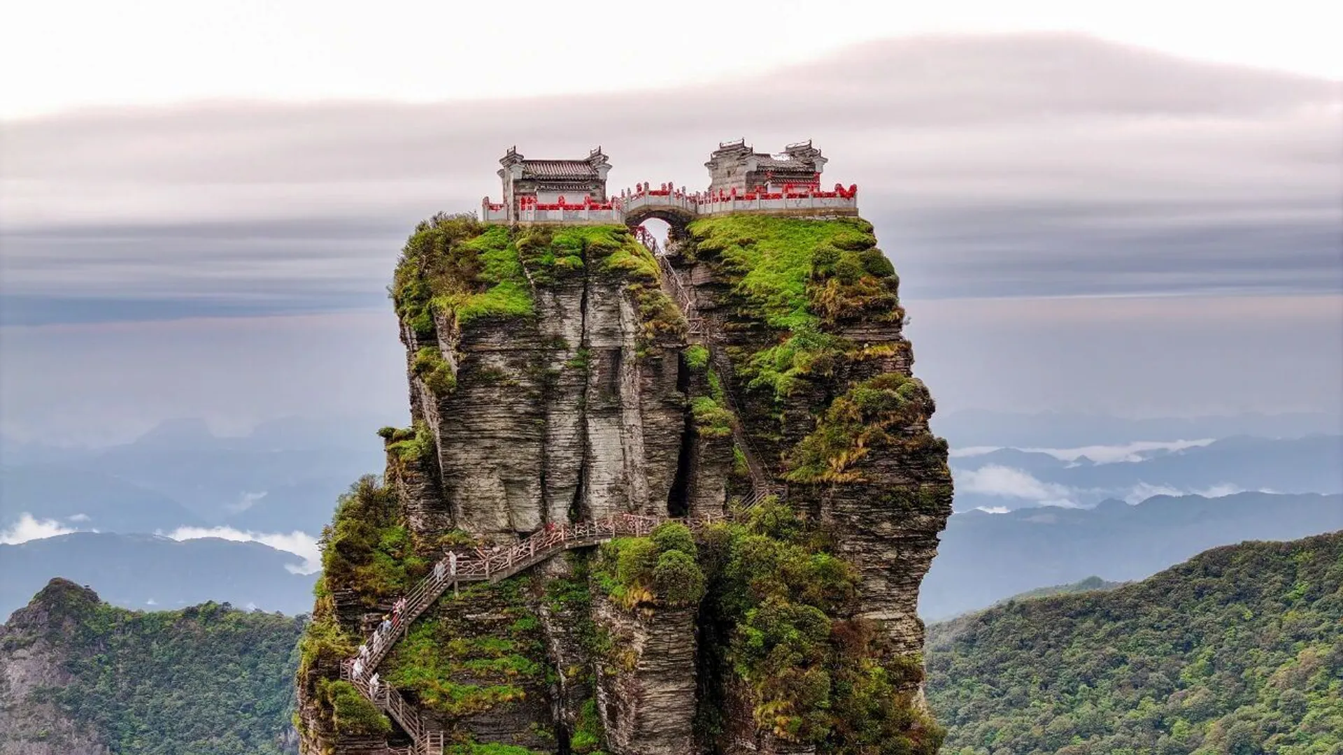 The image shows a dramatic, mountainous landscape with a tall, narrow rock formation. At the top, there are ancient-looking structures, possibly temples or pavilions. A staircase zigzags up the side of the rock, leading to the top. The surrounding area is lush with greenery, and distant mountains are visible under a cloudy sky.