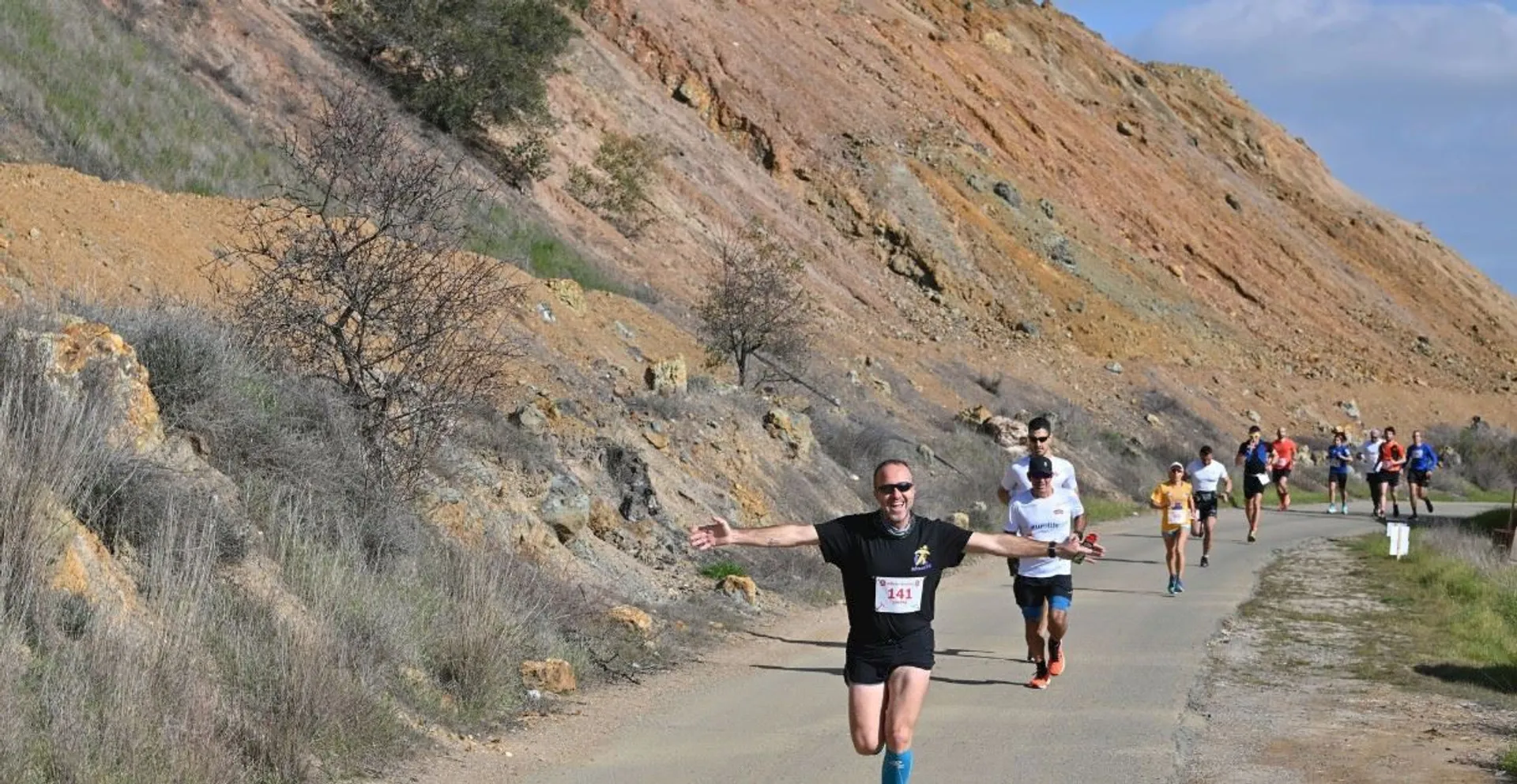 The image shows a group of people running on a paved path in a scenic outdoor area with rugged terrain and rocky hills. One person in the foreground has outstretched arms and is wearing athletic clothing. The setting looks like a race or a running event.