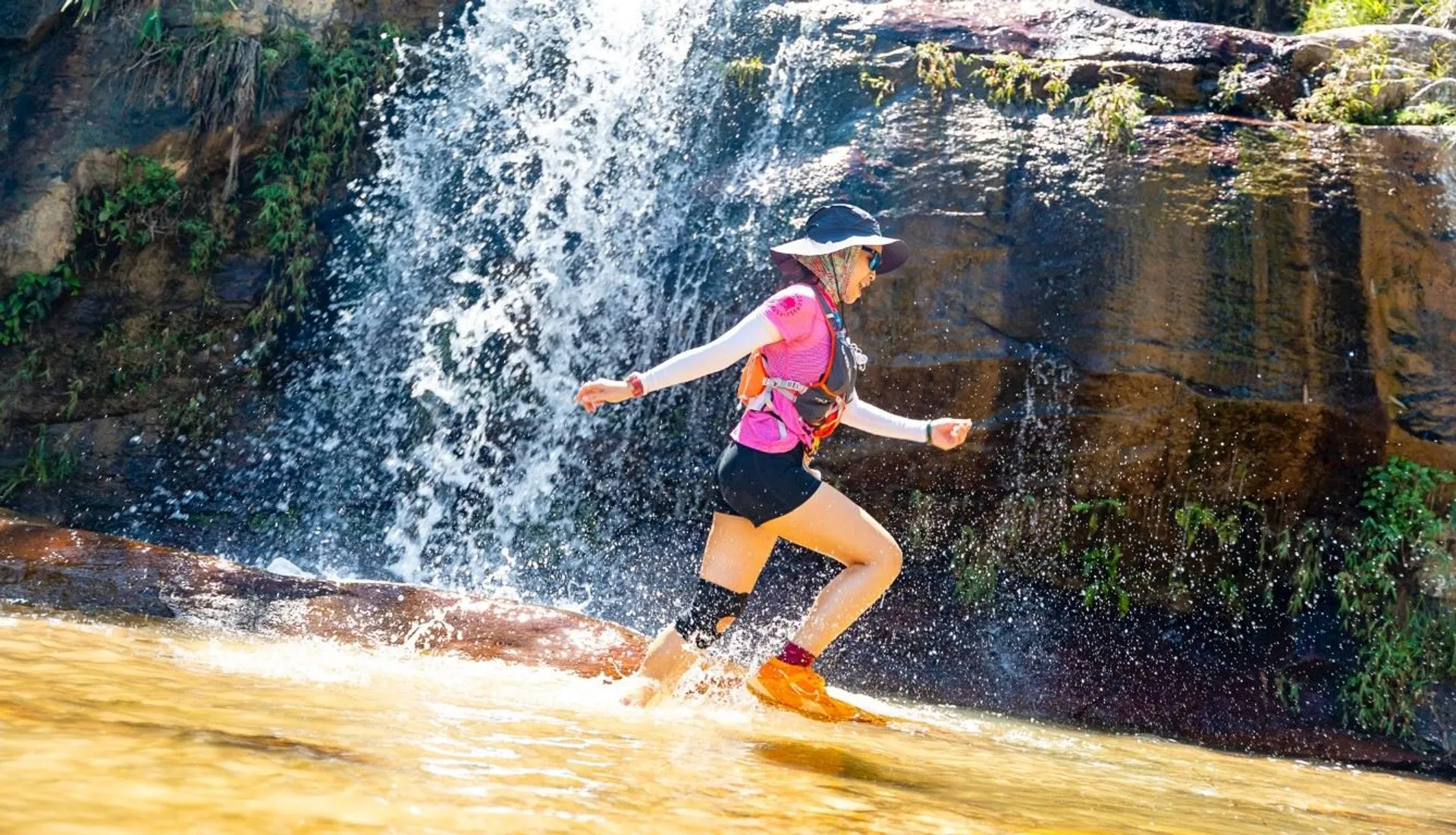 The image shows a person running through shallow water at the base of a waterfall. The individual is wearing a pink shirt, black shorts, a black hat, and has a knee brace. The person also has a hydration pack and is wearing sunglasses. The background features the waterfall and rocks with some vegetation.