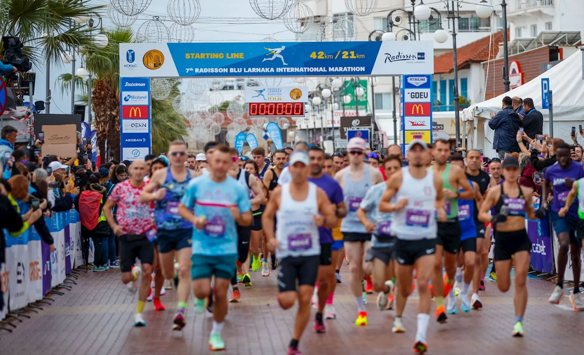 The image shows a group of runners at the starting line of a marathon event. The banner indicates it's the "Radisson Blu Larnaka International Marathon." Spectators are visible along the sides of the course, and various signs and sponsor logos are present. The scene is lively and busy, typical of a race start.