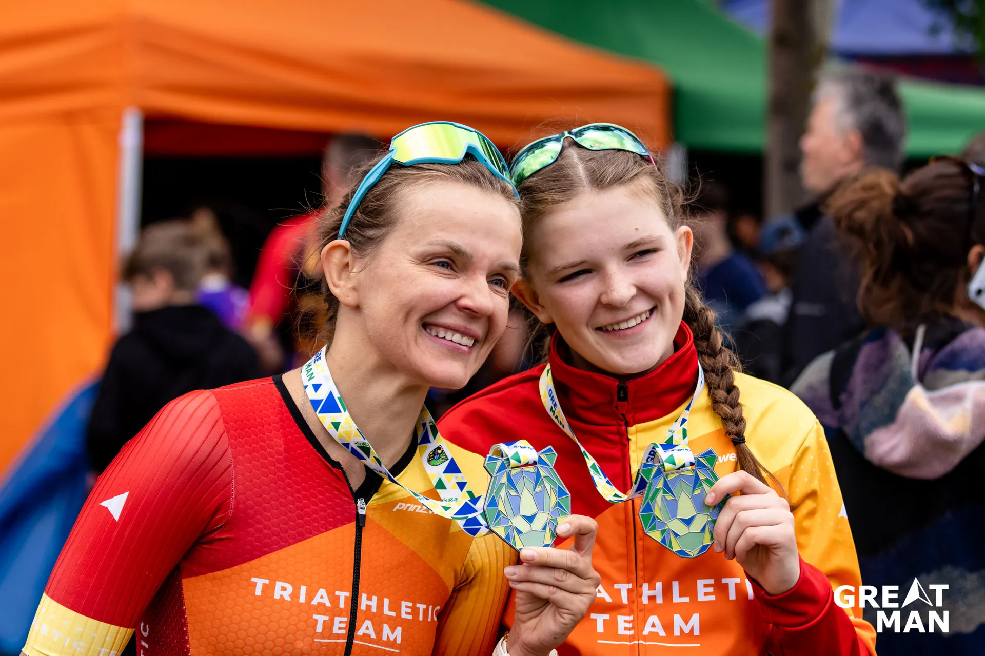 The image shows two people posing with medals, likely after completing a sporting event. They are both wearing athletic gear, possibly from a triathlon team, and there are tents and other people in the background.