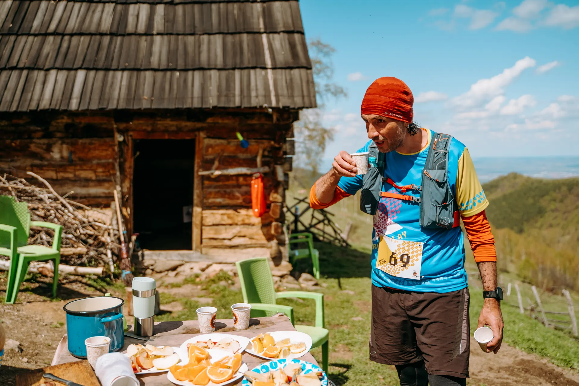 The image shows a person wearing a race bib and outdoor running gear, including a red head covering and a blue shirt, drinking from a cup. They are standing near a small wooden cabin with a slanted roof. In front of them is a table with refreshments, including sliced oranges and cups. There are also green chairs around the area, and the scene is set in a mountainous or hilly environment with a clear blue sky.