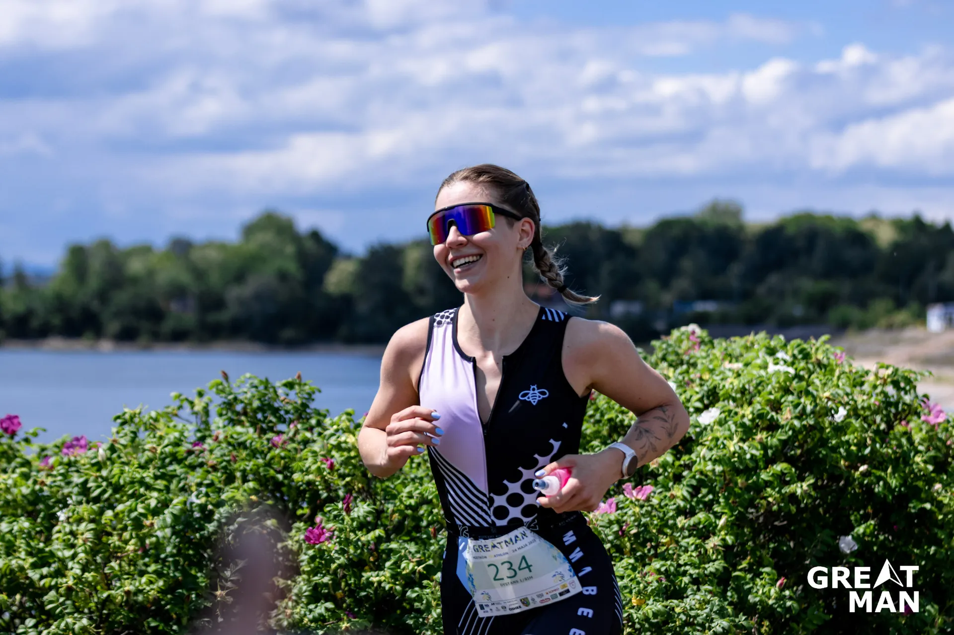The image shows a person running outdoors, wearing athletic attire that includes a sleeveless shirt with patterns, sunglasses, and a race bib numbered 234. The person is smiling and carrying something pink in their hand. The background features greenery and a body of water, suggesting a scenic location, possibly by a lake or a river with a clear sky.