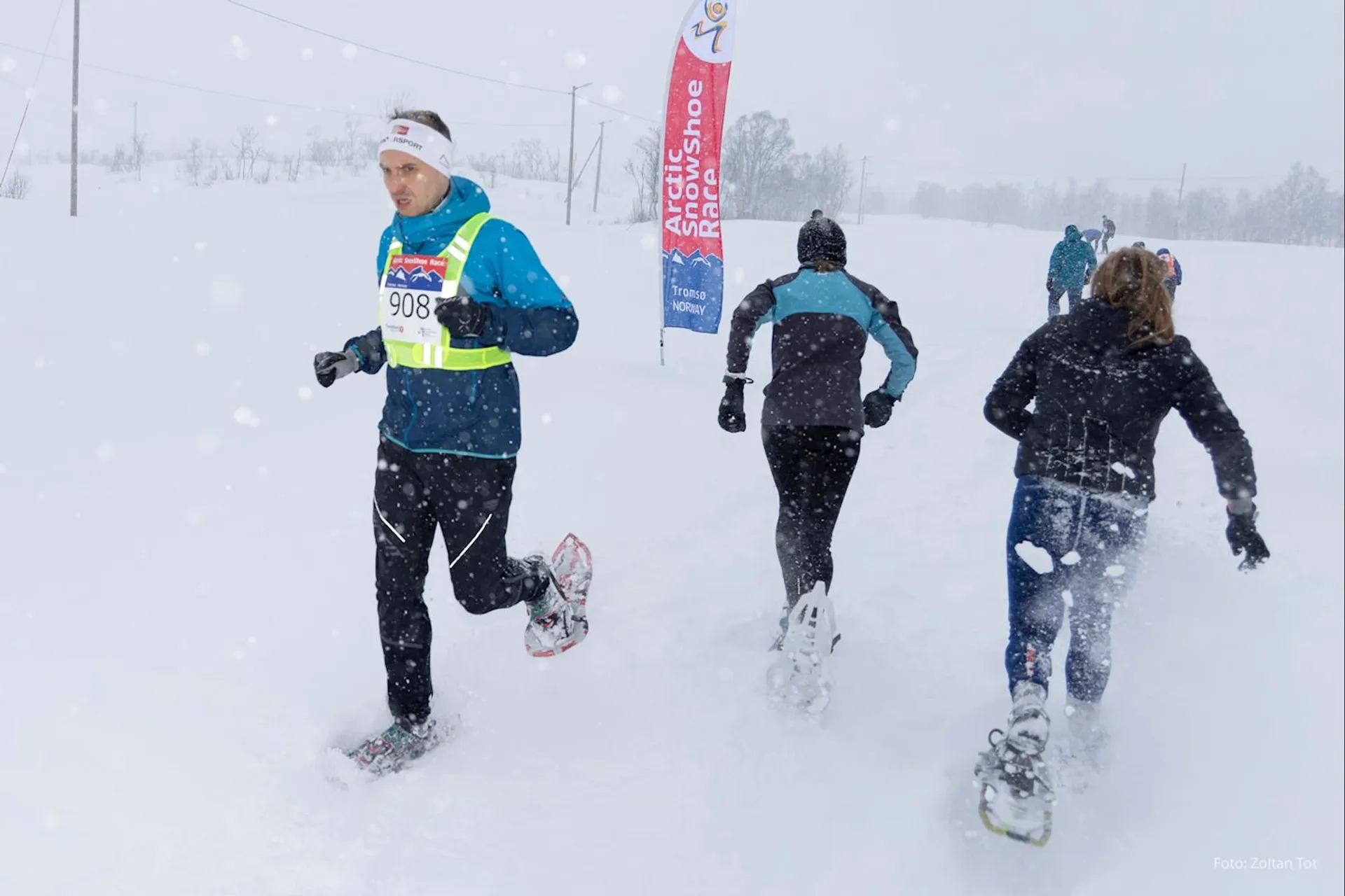 This image shows a group of people participating in a snowshoeing event or race. They are outdoors and it appears to be actively snowing, given the snowflakes visible in the air and the snow-covered ground. The individual marked with the race number 908 is wearing a winter sporting outfit, including a hat and gloves, suitable for cold weather activities. The participants are wearing snowshoes, which are specialized footwear for walking over snow. There are banners along the course, suggesting that this is an organized event. Everyone looks focused and determined, pushing through the snowy conditions.