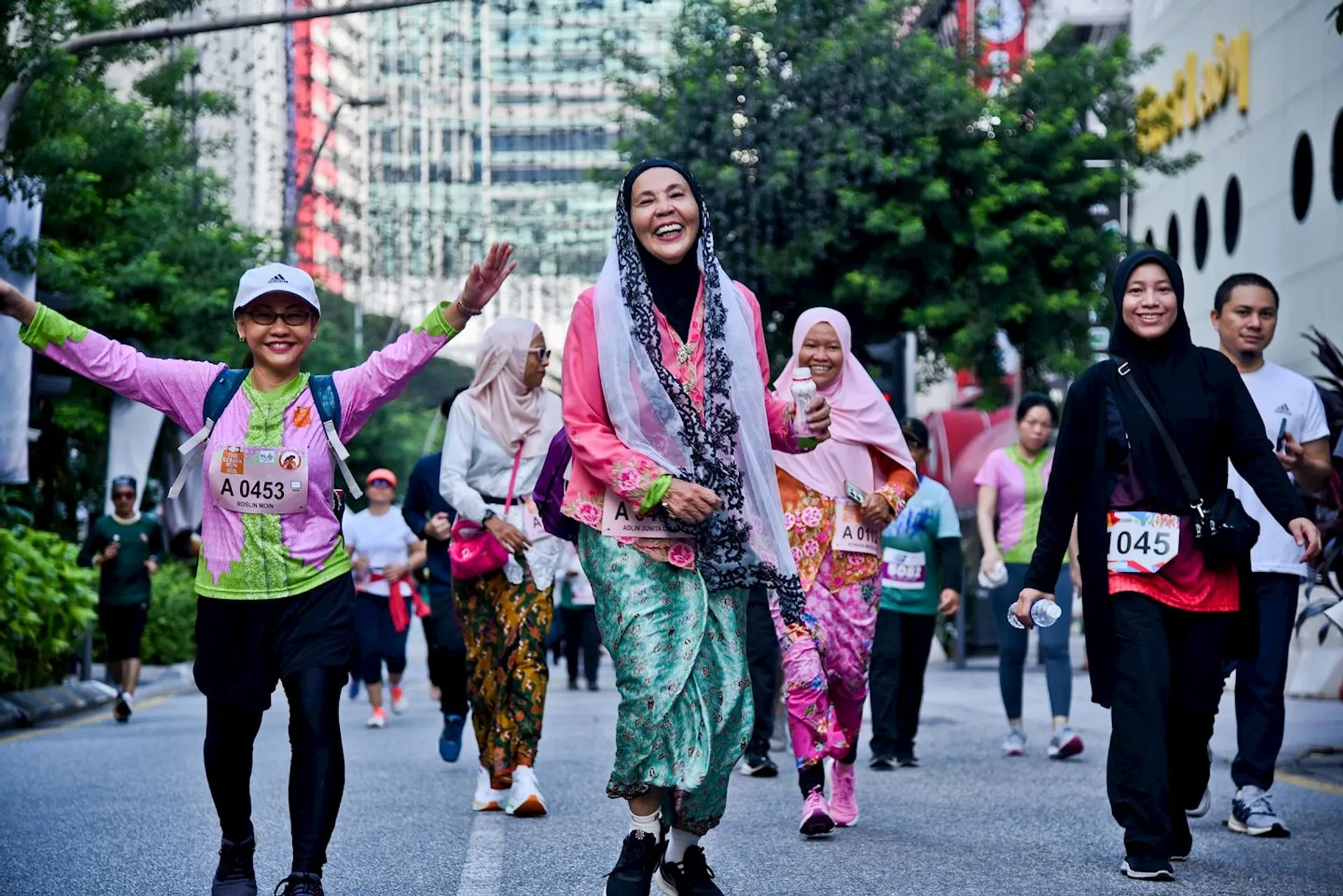 The image shows a group of people participating in an outdoor event, like a run or walk, on a street. The participants are wearing comfortable clothing and bib numbers. They appear to be enjoying the event, as some are smiling and one person has their arms raised. The setting includes urban and natural elements in the background.