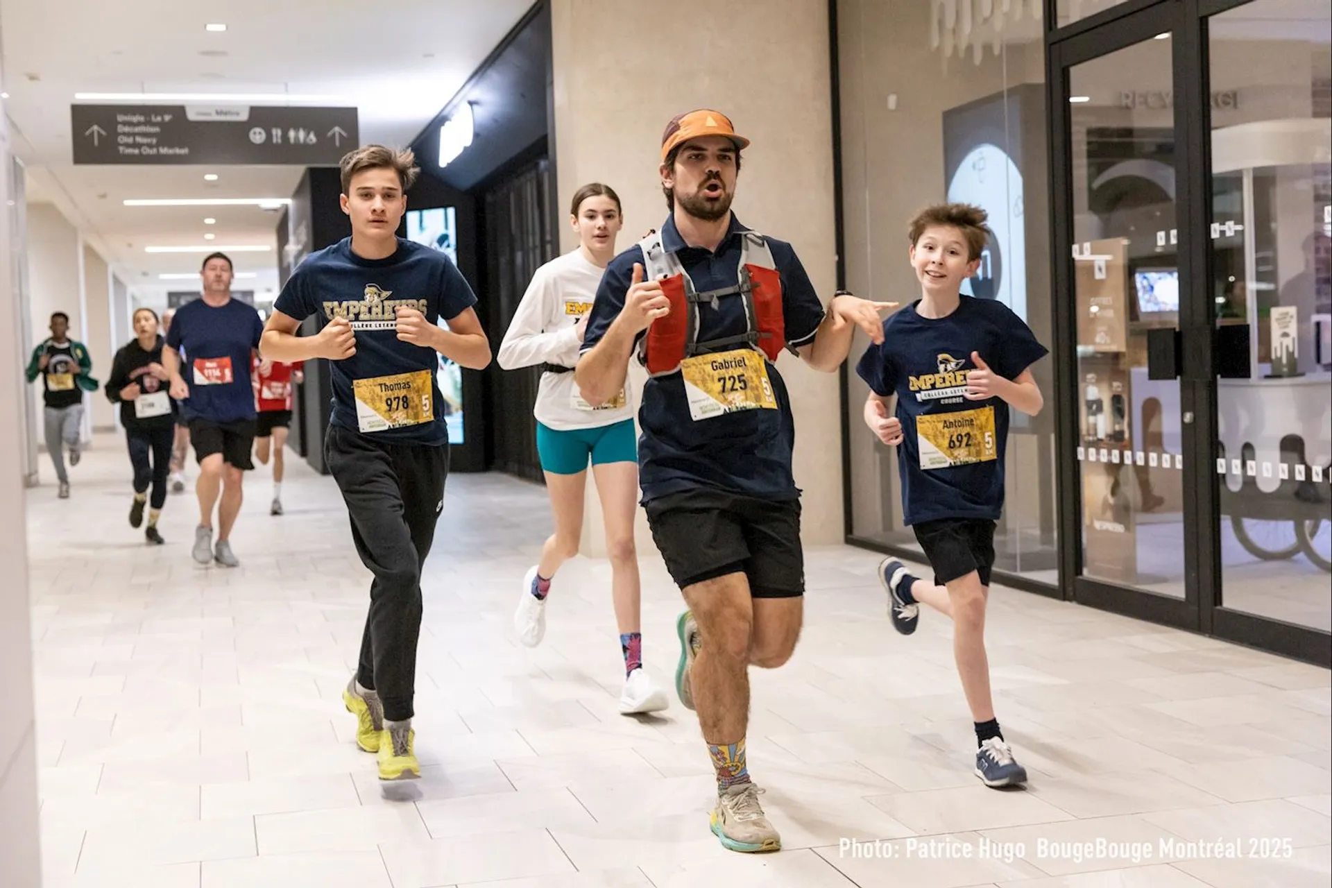 The image shows a group of people participating in an indoor running event. They are running through a shopping mall or similar setting. The participants are wearing athletic clothing with bibs that likely display their numbers and possibly the name of the event. There are signs and store fronts visible in the background.