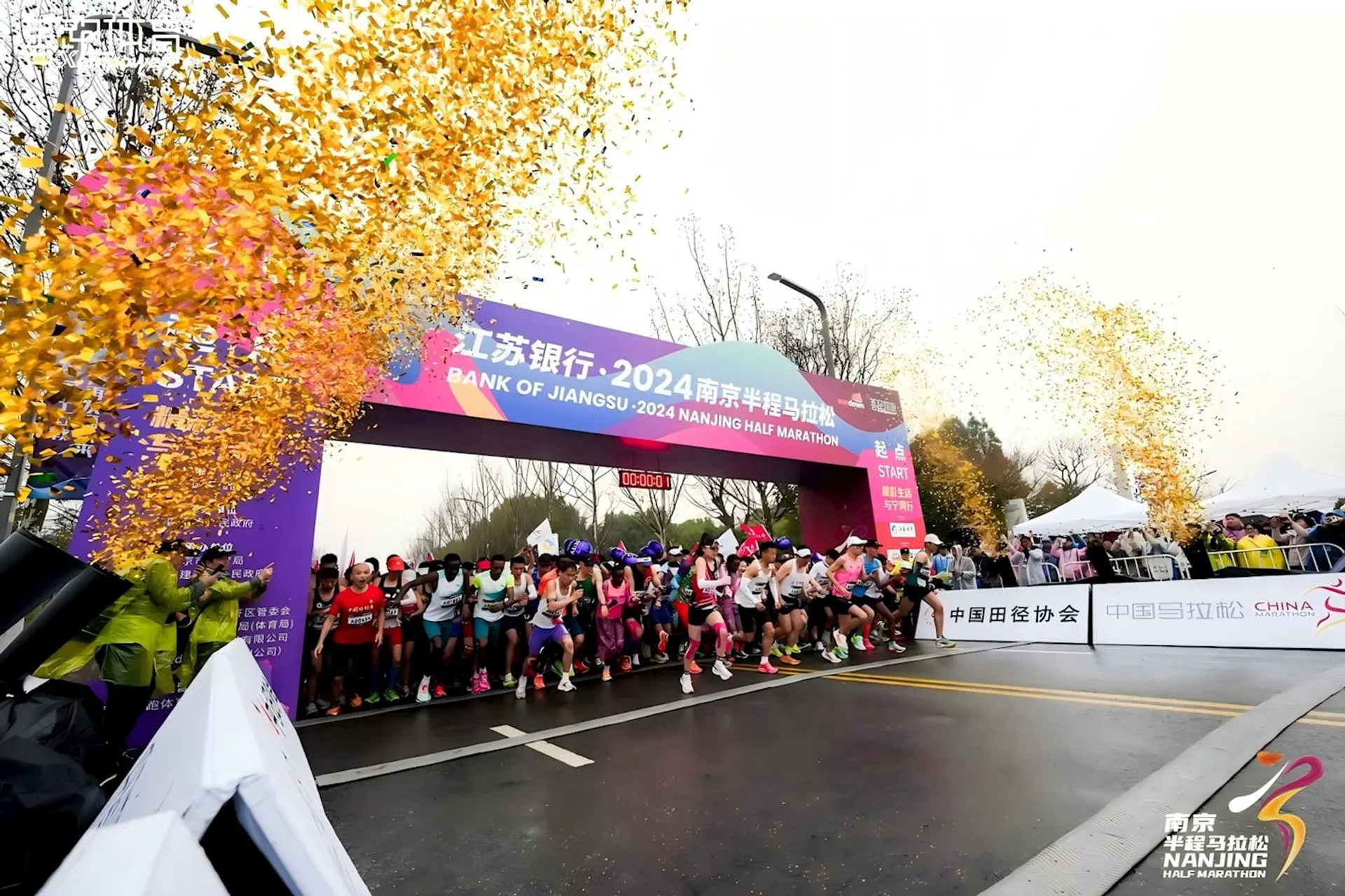 The image shows the start of the "Bank of Jiangsu 2024 Nanjing Half Marathon." Runners are lined up at the starting line, and confetti is falling from above. There are banners and logos related to the event, indicating sponsorship and location.