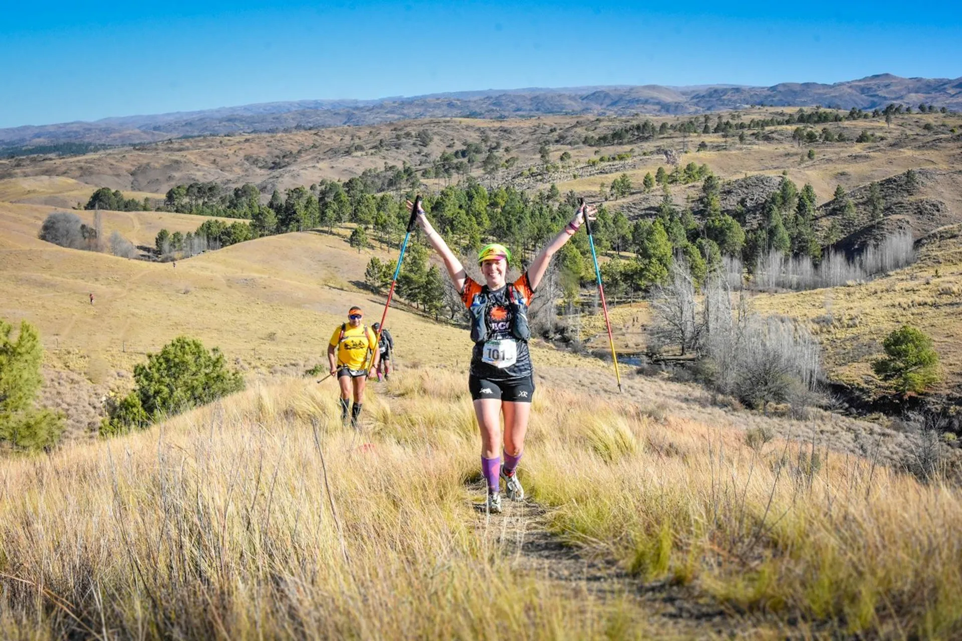 The image shows a person running or hiking on a trail in a grassy, hilly landscape. They appear to be participating in a trail race, wearing athletic gear, with a race bib, and holding trekking poles. Other participants are visible in the background. The environment is an open, scenic area with rolling hills.